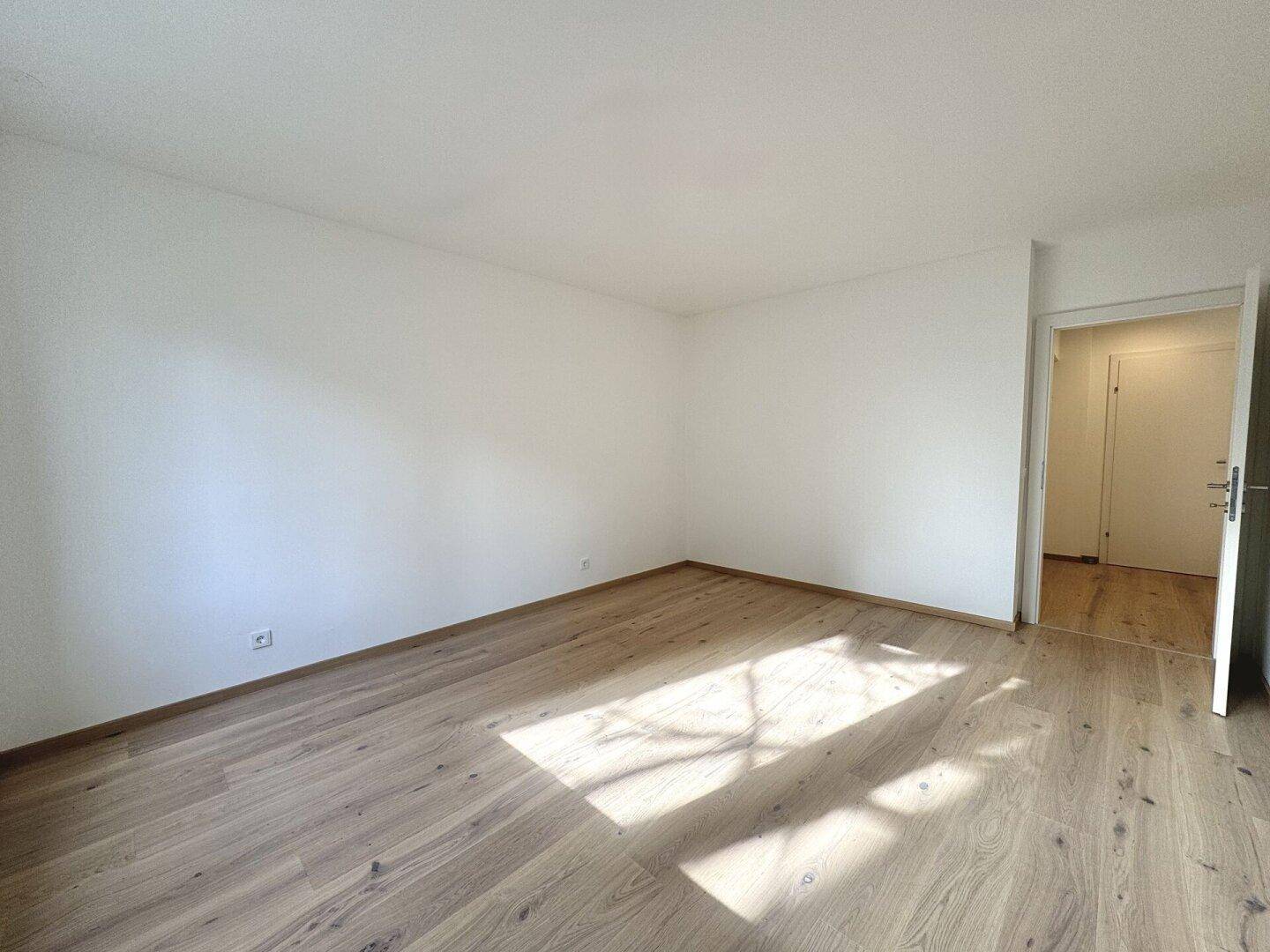 Leerer Raum mit sonnigem Holzparkettboden und weiß gestrichenen Wänden, einladend und hell.