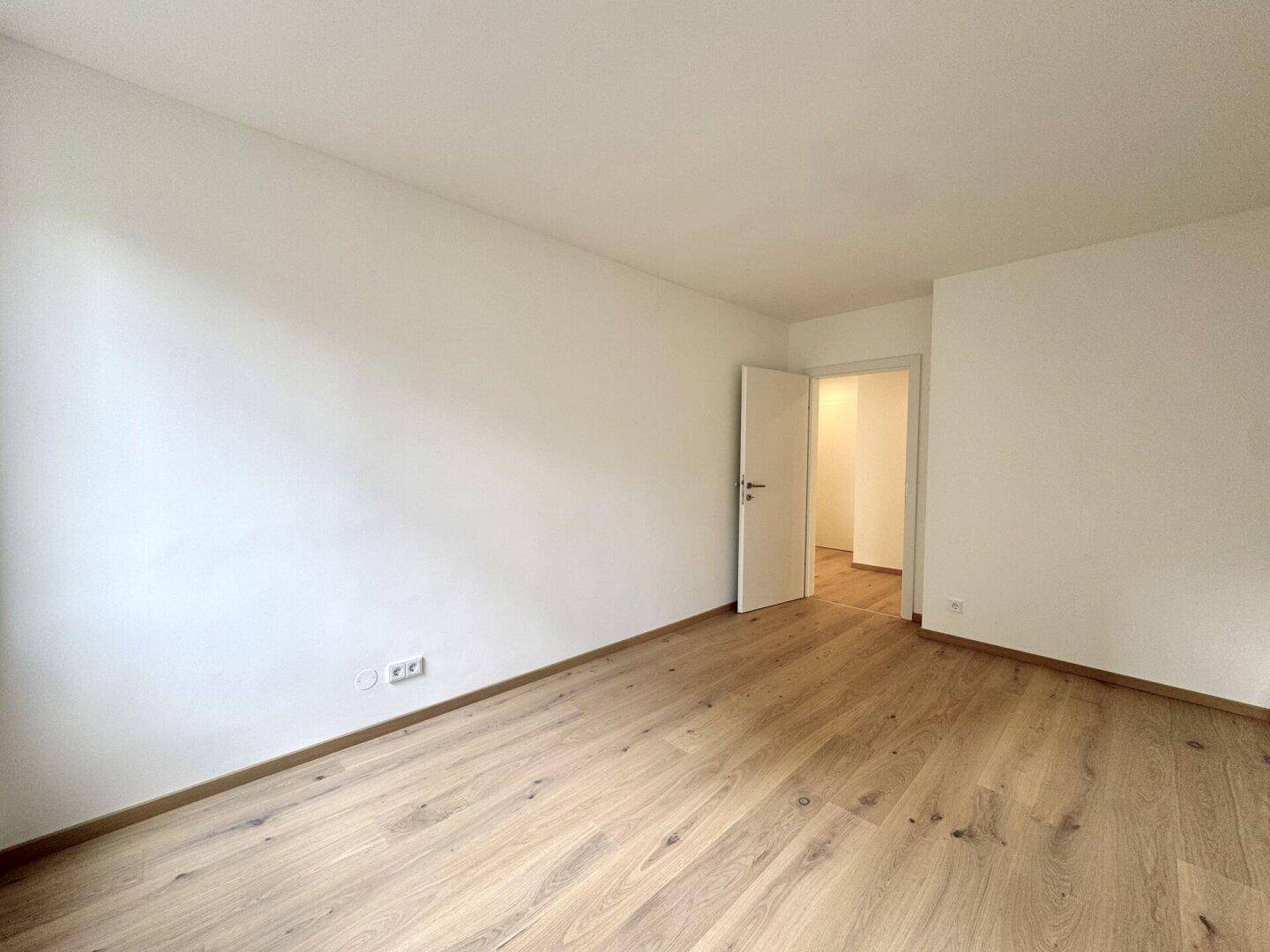 Leerer Raum mit Holzparkettboden und offener Tür zu einem angrenzenden Bereich, hell und einladend.