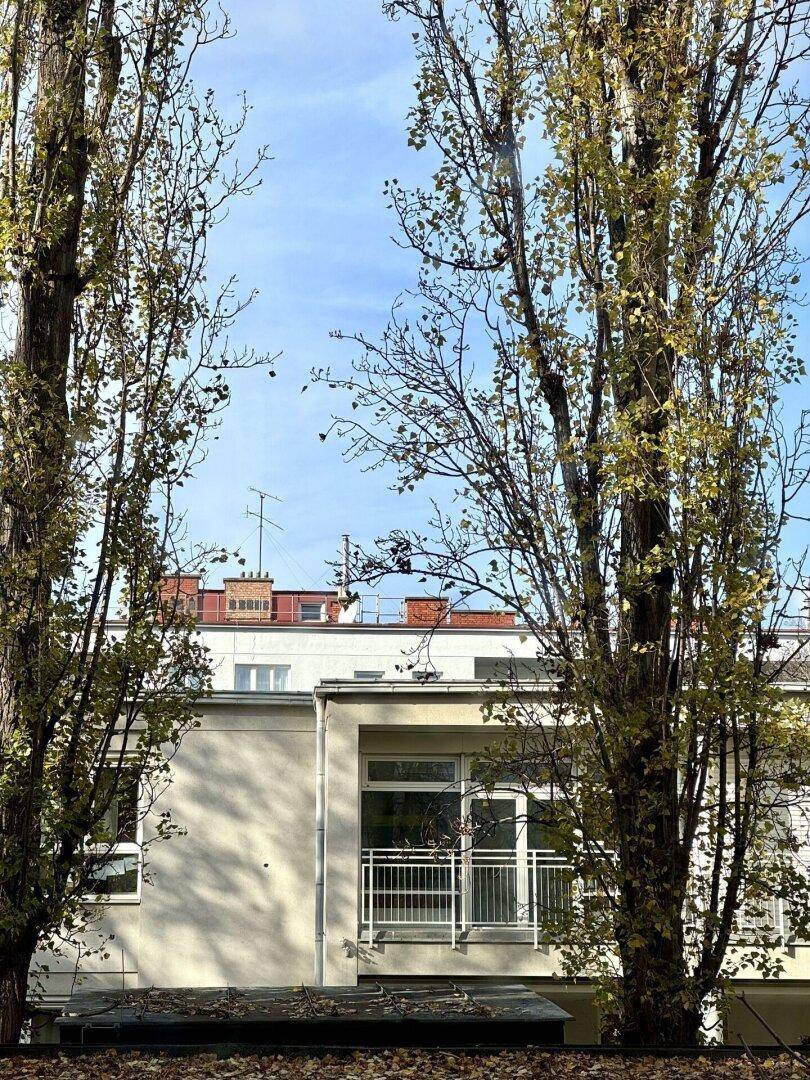 Fassade des Gebäudes mit kleinem Balkon, umgeben von hohen Bäumen und blauem Himmel.