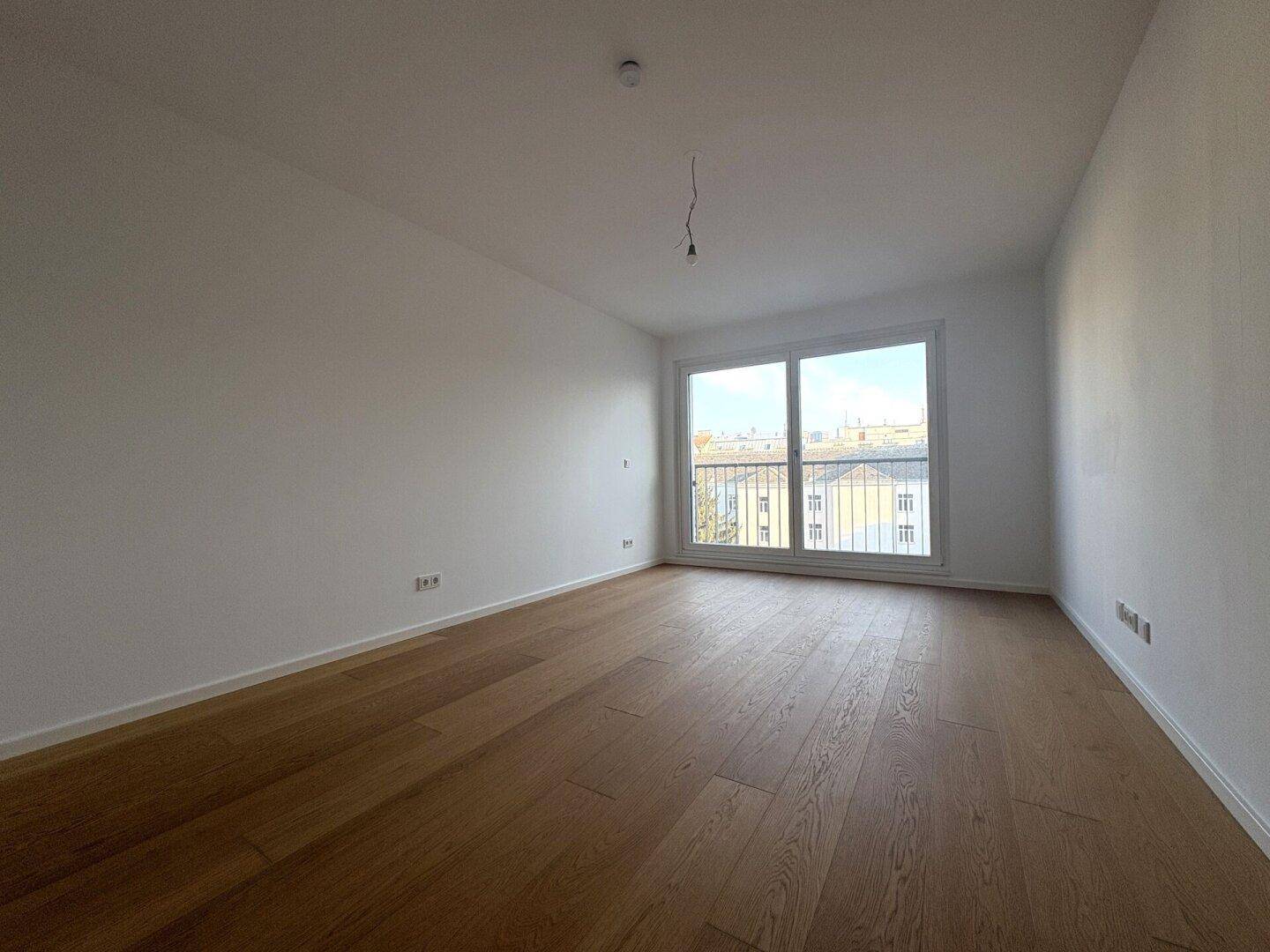 Leeres Schlafzimmer mit Holzboden und großem Fenster, das viel Licht hereinlässt.