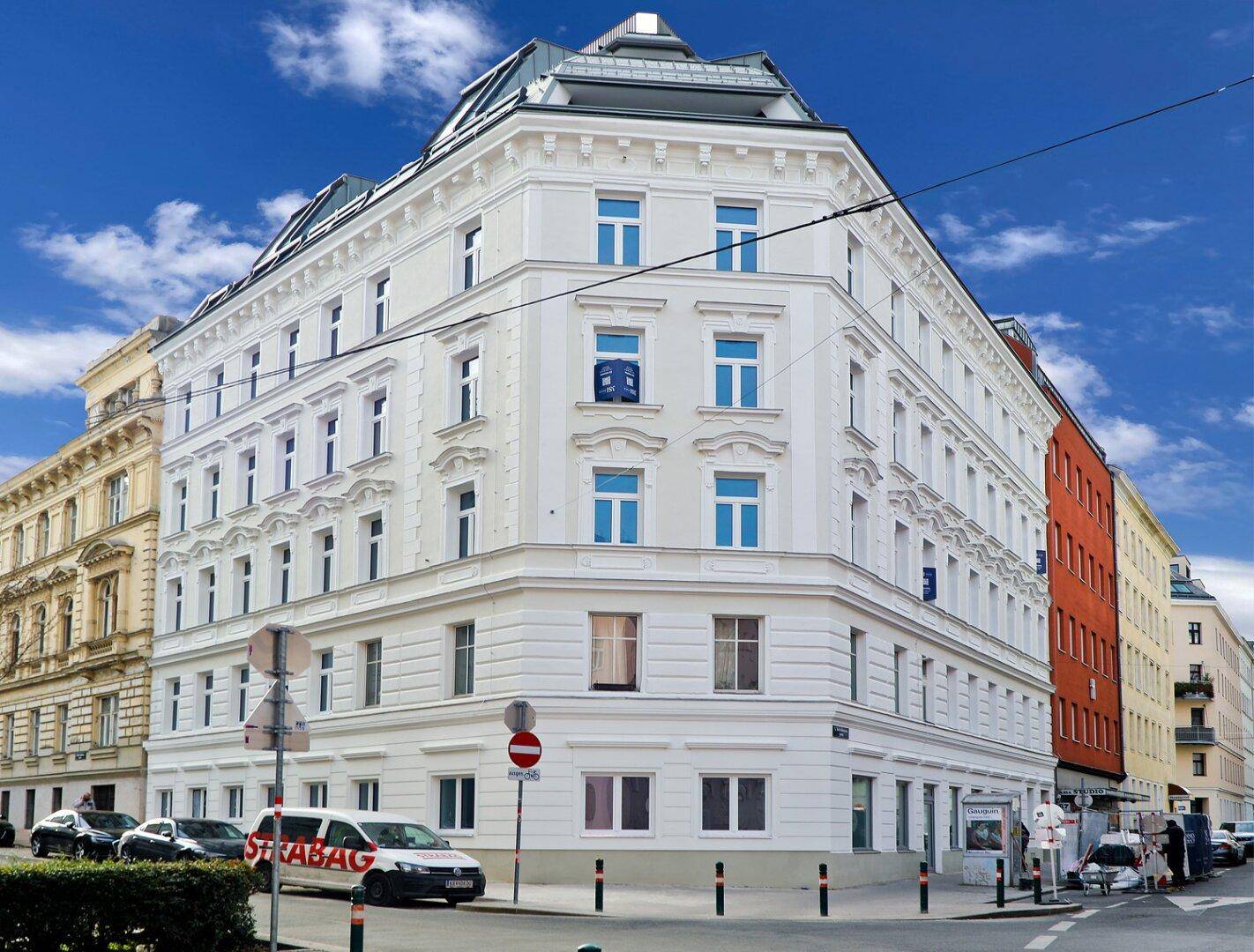 Historisches Eckgebäude mit weißer Fassade und blauen Fenstern an einer belebten Straße.