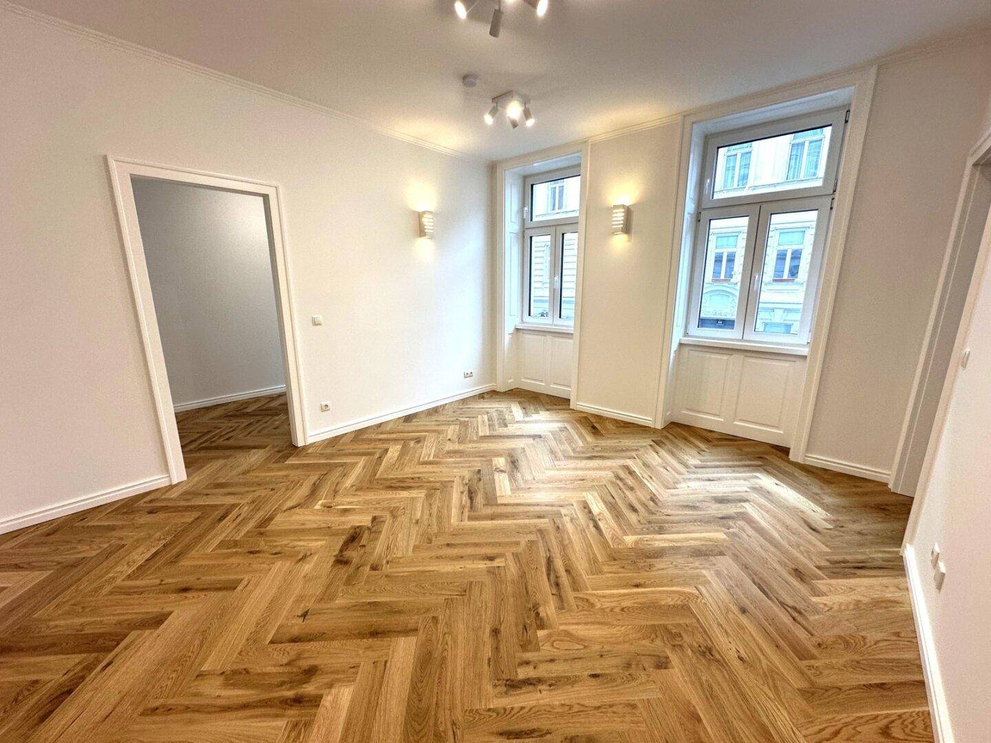 Großes Wohnzimmer mit Fischgrätparkett, hohen Decken und zwei großen Fenstern.