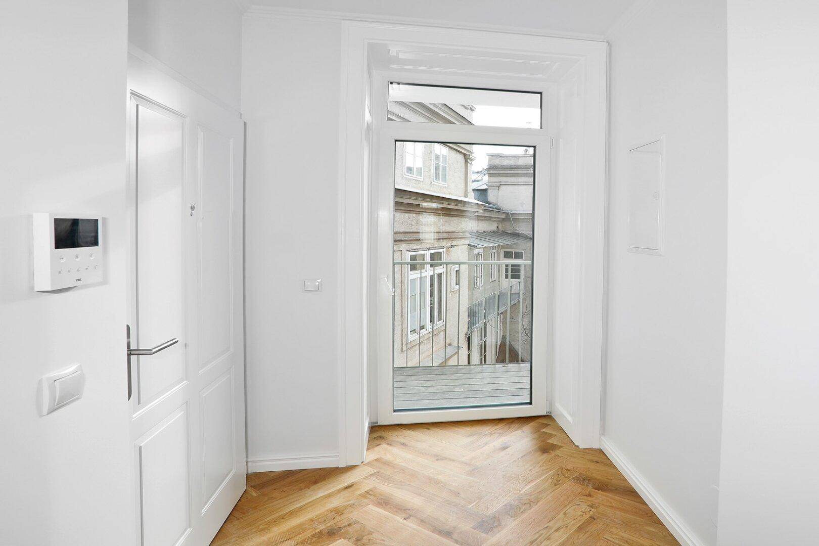 Heller Flur mit Fischgrätparkett und Blick auf die umliegenden Gebäude durch einen französischen Balkon.