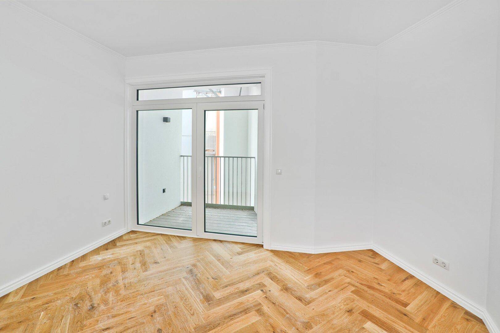 Helles Zimmer mit Fischgrätparkett und Zugang zu einem Balkon durch eine große Glastür.