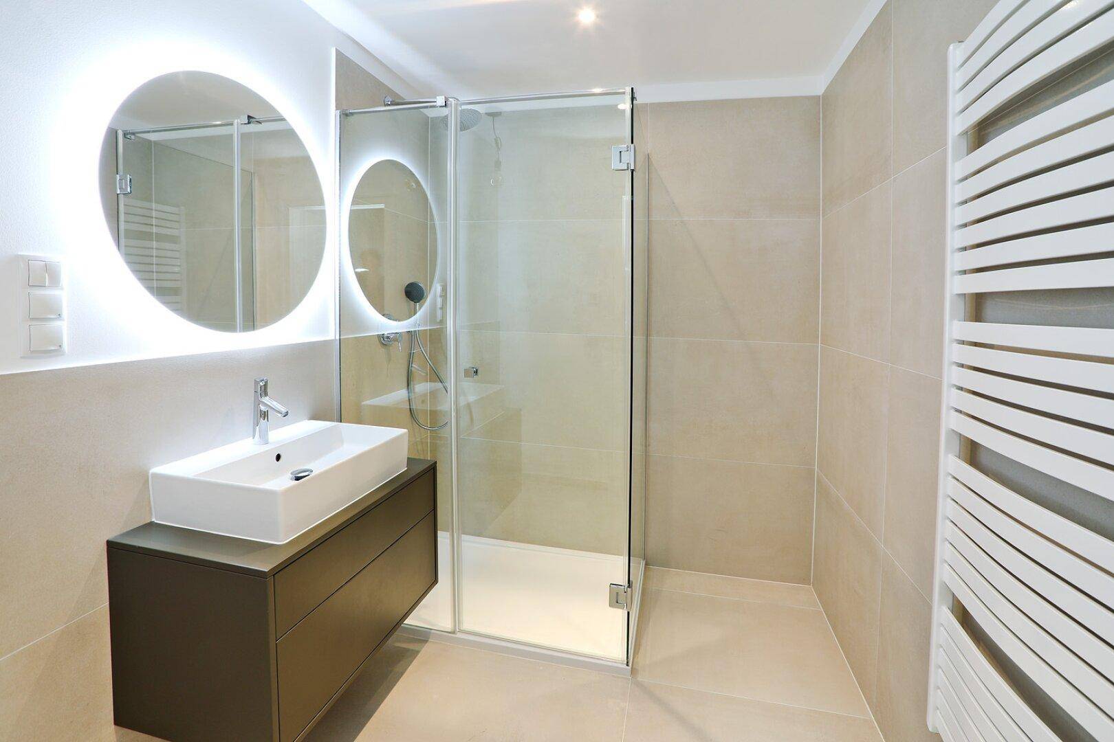 Modernes Badezimmer mit begehbarer Dusche, beleuchtetem Spiegel und beheiztem Handtuchhalter.