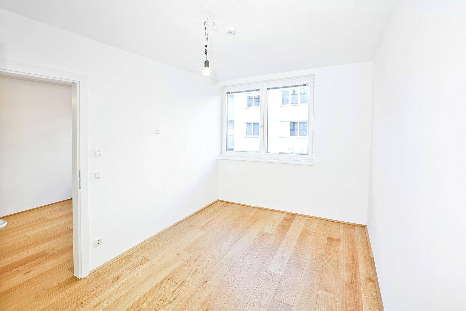 Helles Zimmer mit großen Fenstern, weiß gestrichenen Wänden und Eichenparkettboden.