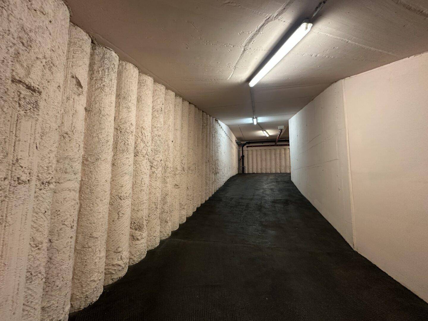 Abfallende Rampe zur Tiefgarage mit strukturierten Betonwänden und Deckenbeleuchtung.