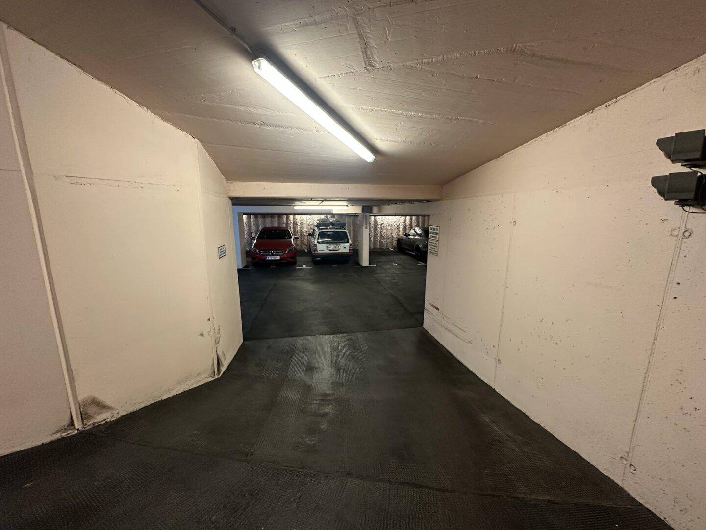 Einfahrt zur Tiefgarage mit Betonwänden und Deckenbeleuchtung, die zu weiteren Parkplätzen führt.