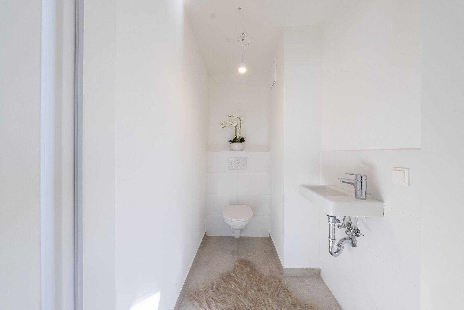 Modernes Gäste-WC mit Wand-WC, kleinem Waschbecken und hellem Fliesenboden.
