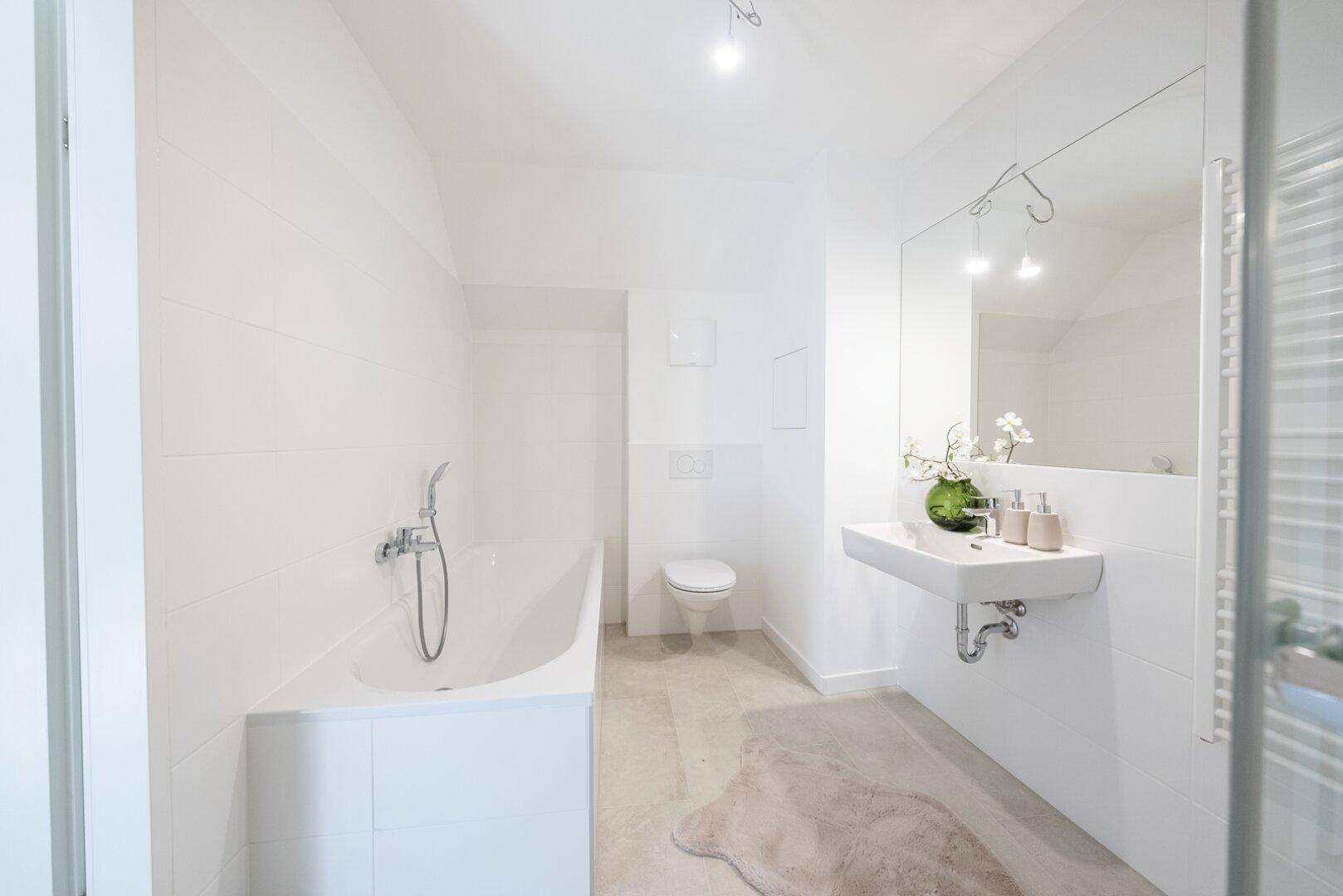 Modernes Badezimmer mit Badewanne, Wand-WC und Waschbecken, alles in hellen Farbtönen gehalten.