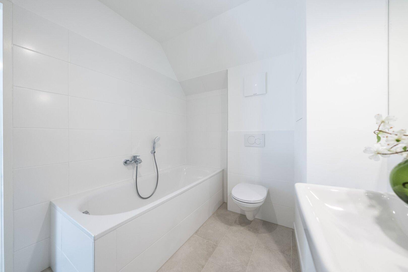 Detailansicht der Badewanne und des Wand-WCs in einem modernen, hell gefliesten Badezimmer.