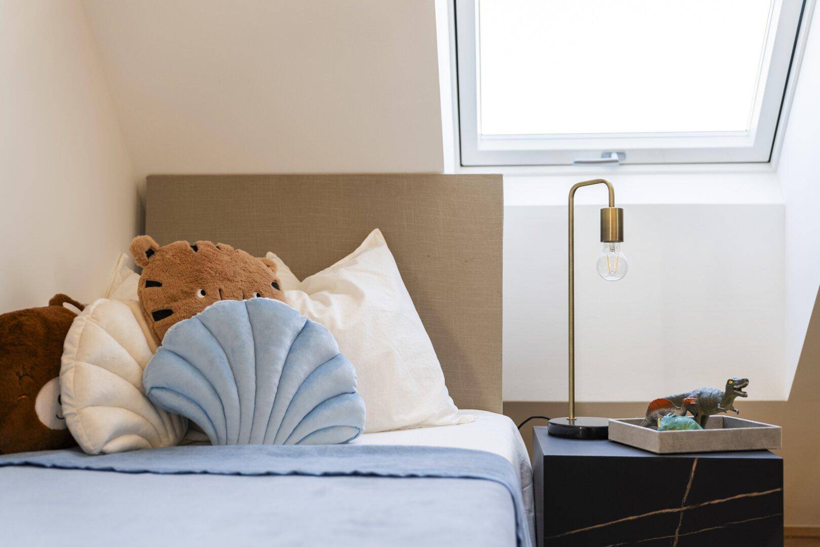Kinderzimmer mit Einzelbett, Dachfenster und Nachttisch mit moderner Lampe.