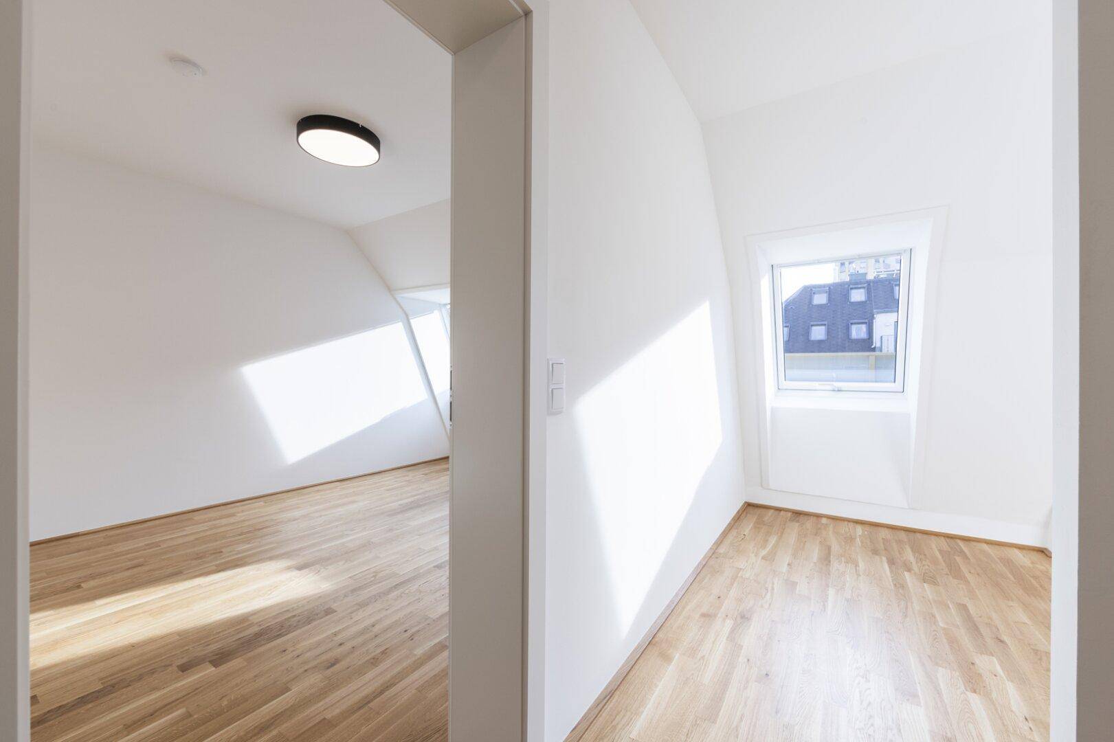 Leeres Zimmer mit Parkettboden, Dachschräge und einem Fenster, das viel Tageslicht hereinlässt.
