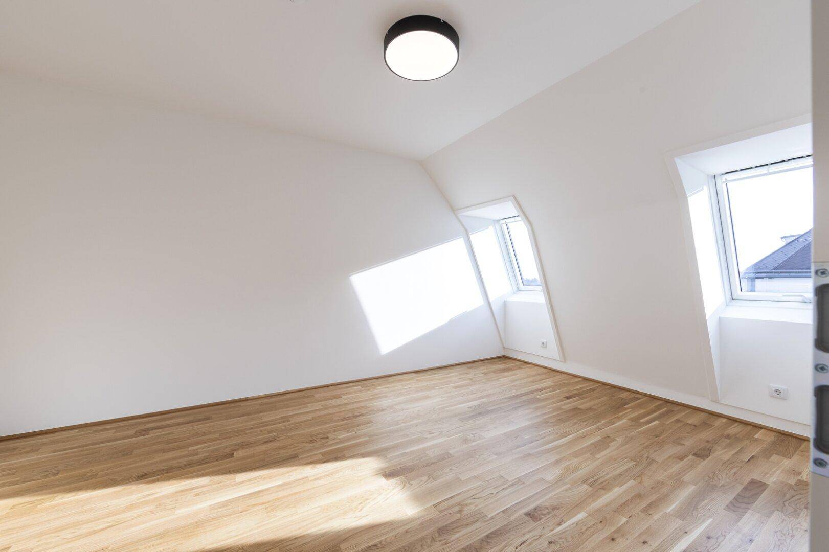 Helles Zimmer mit Parkettboden, weißen Wänden und mehreren Fenstern unter der Dachschräge.
