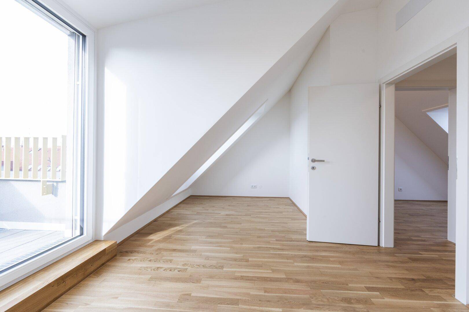 Heller Raum mit Holzparkett, schräger Decke und großem Fenster mit Zugang zur Terrasse.