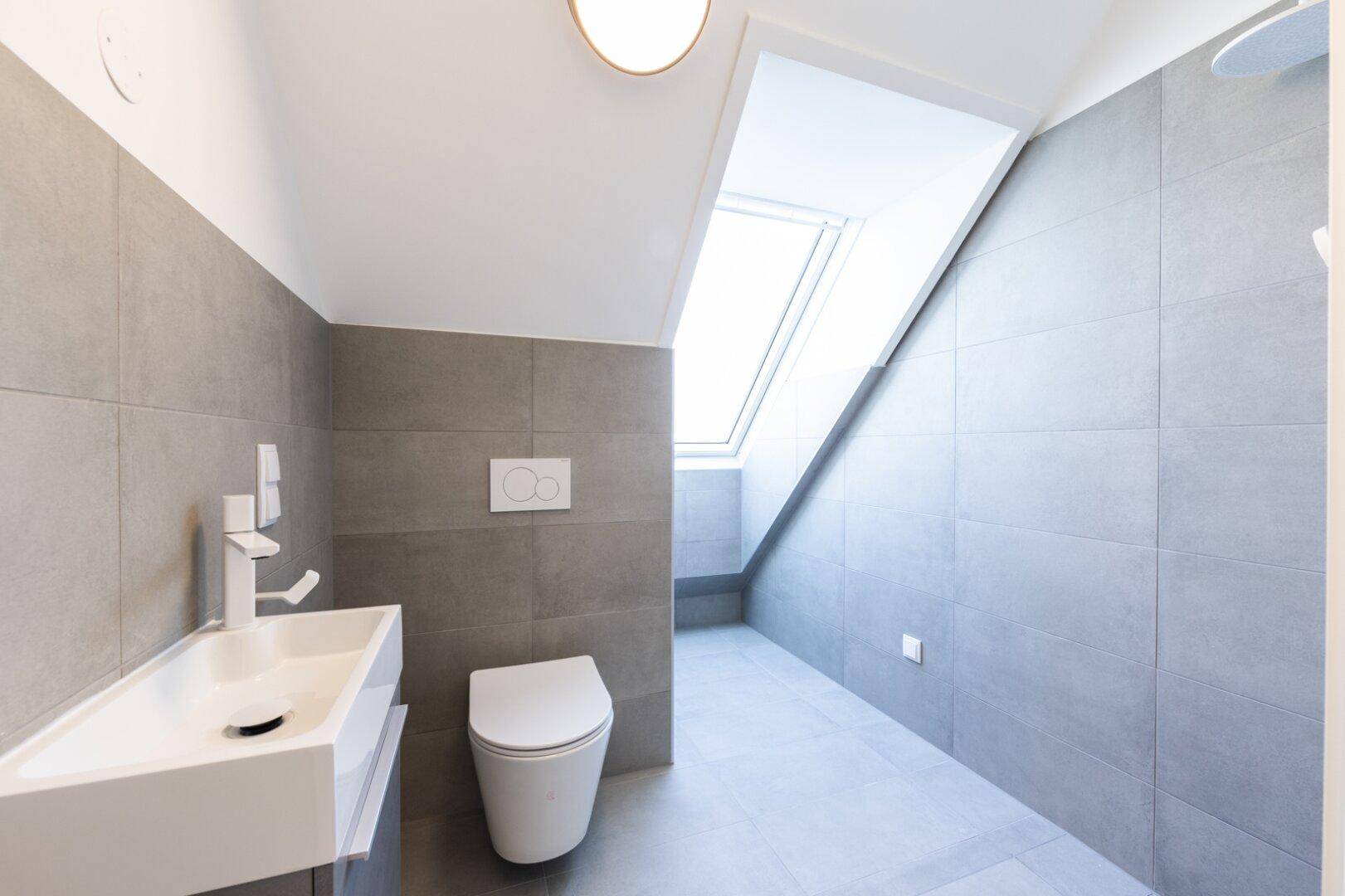 Modernes Badezimmer mit grauen Fliesen, wandhängendem WC und Dachfenster für natürliches Licht.