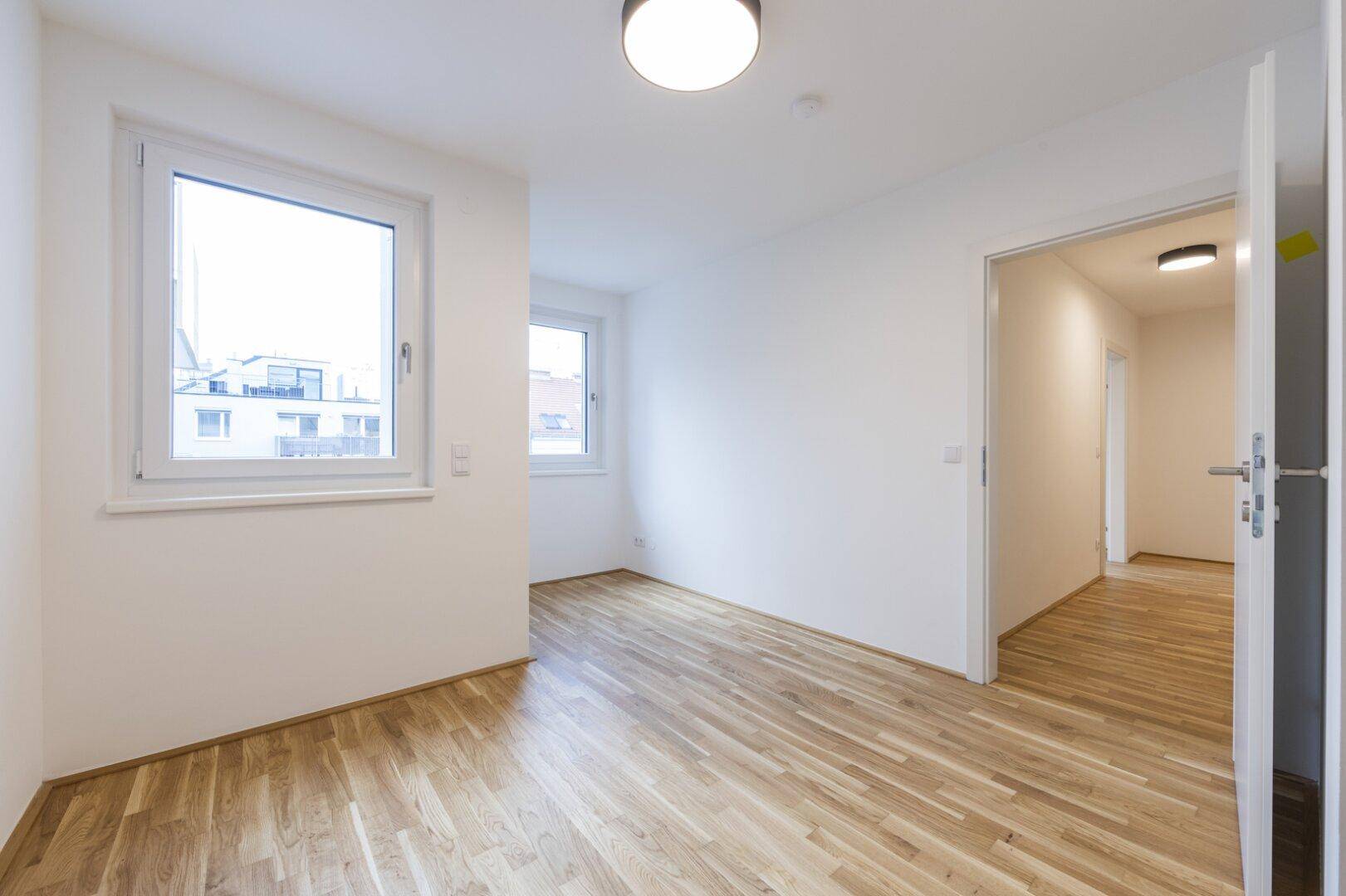 Helles Zimmer mit Parkettboden und zwei Fenstern, mit Blick in den angrenzenden Flur.