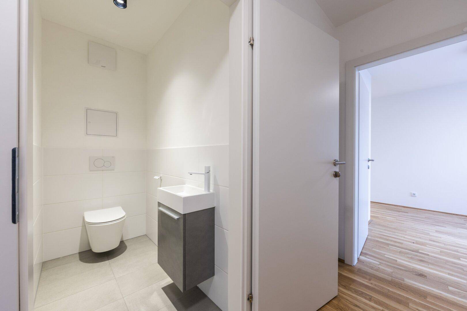 Modernes Gäste-WC mit wandhängender Toilette, kleinem Waschbecken und hellen Fliesen.
