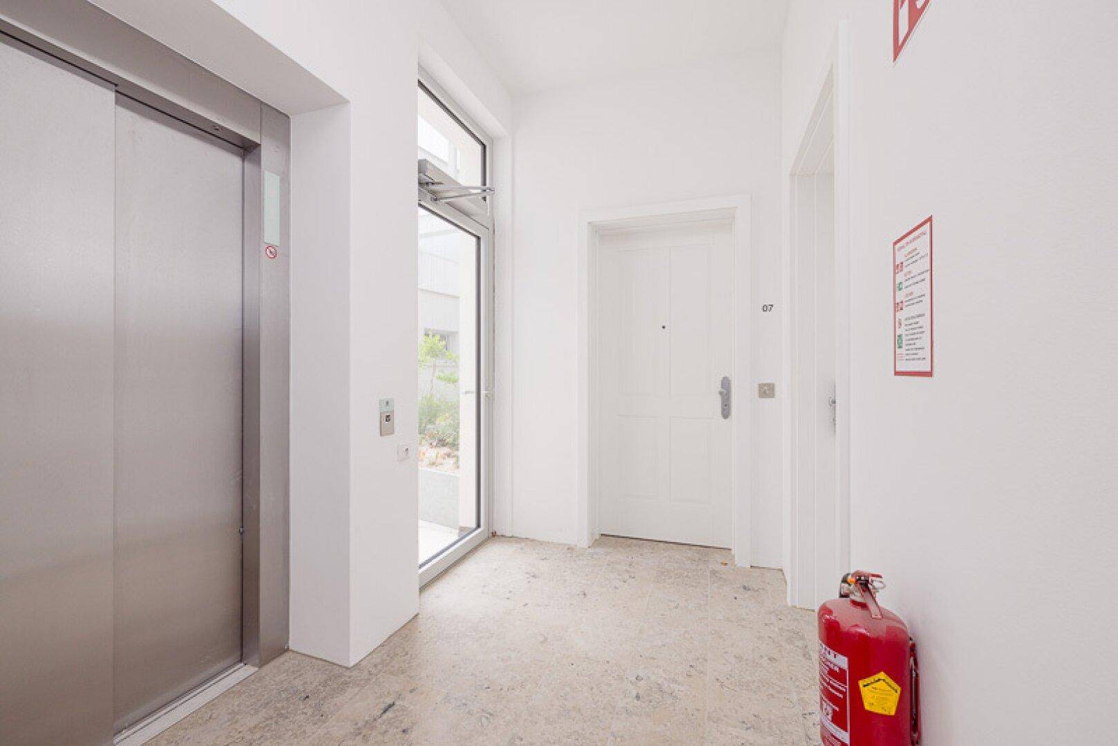 Heller Eingangsbereich mit modernem Aufzug, Wohnungstür und Brandschutzgerät, funktional gestaltet.