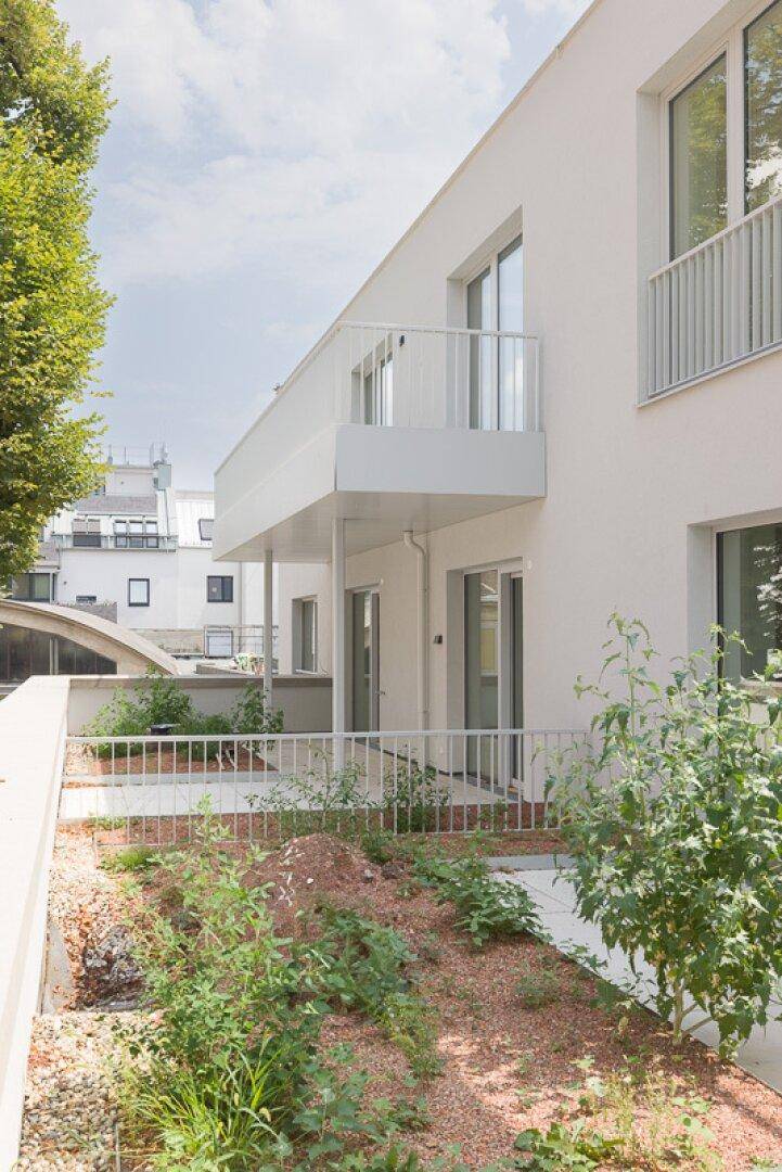 Moderner weißer Gebäudeteil mit kleinem Gartenbereich und Balkon im Freien, frisch bepflanzt.