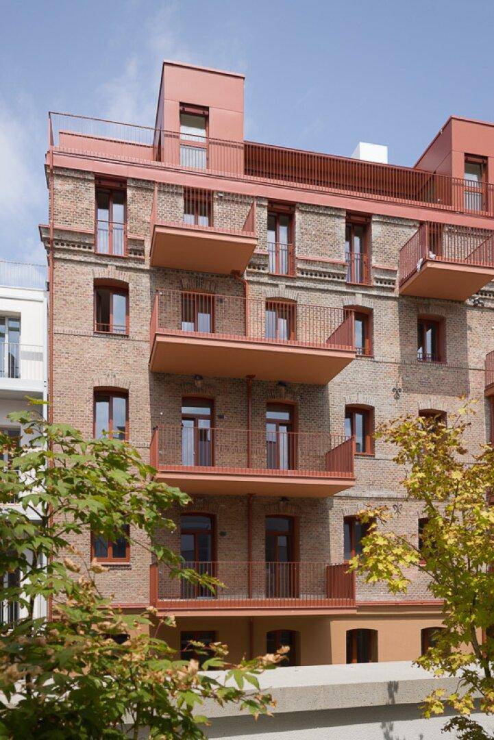 Fassade eines modernen Wohngebäudes mit Ziegelmauerwerk, roten Balkonen und Dachaufbauten.