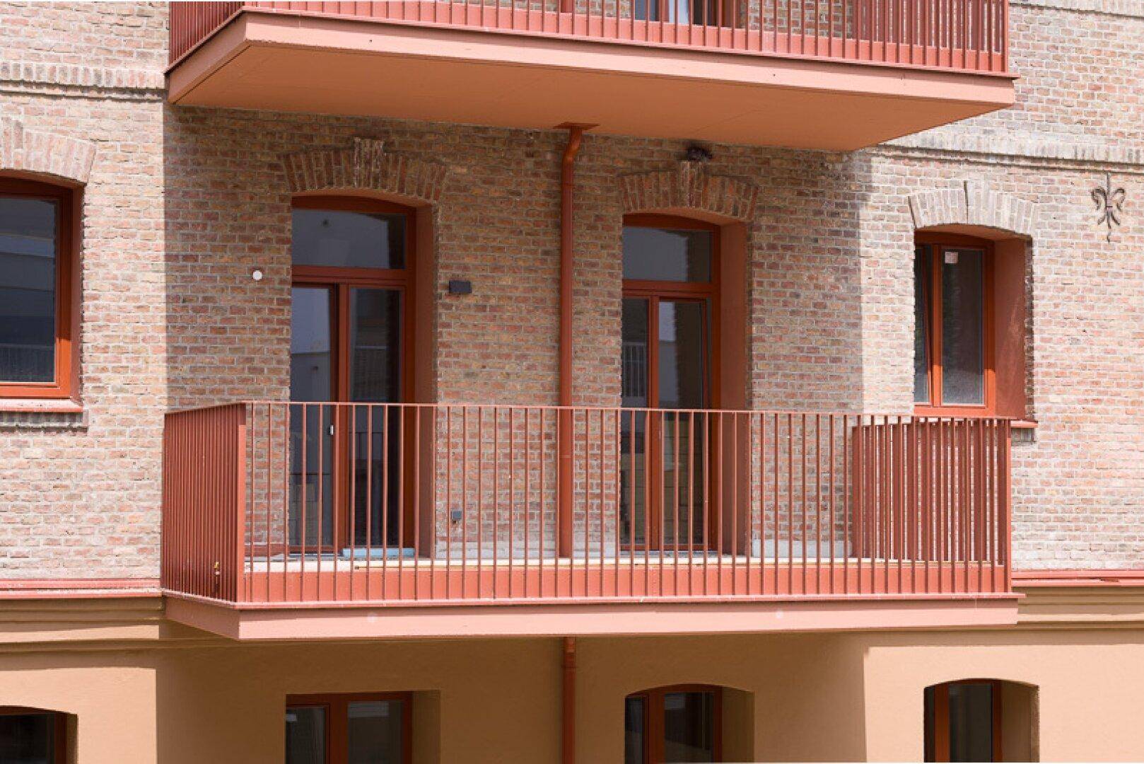 Großer roter Balkon mit Metallgeländer an einer Backsteinwand mit Rundbogenfenstern, einladend gestaltet.