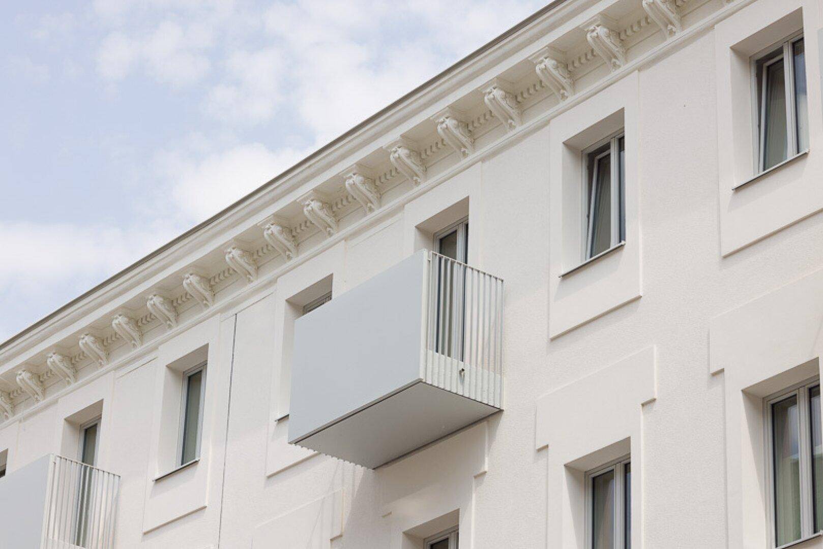Weißer Balkon mit minimalistischem Geländer an einer historischen Fassade mit Stuckverzierungen.
