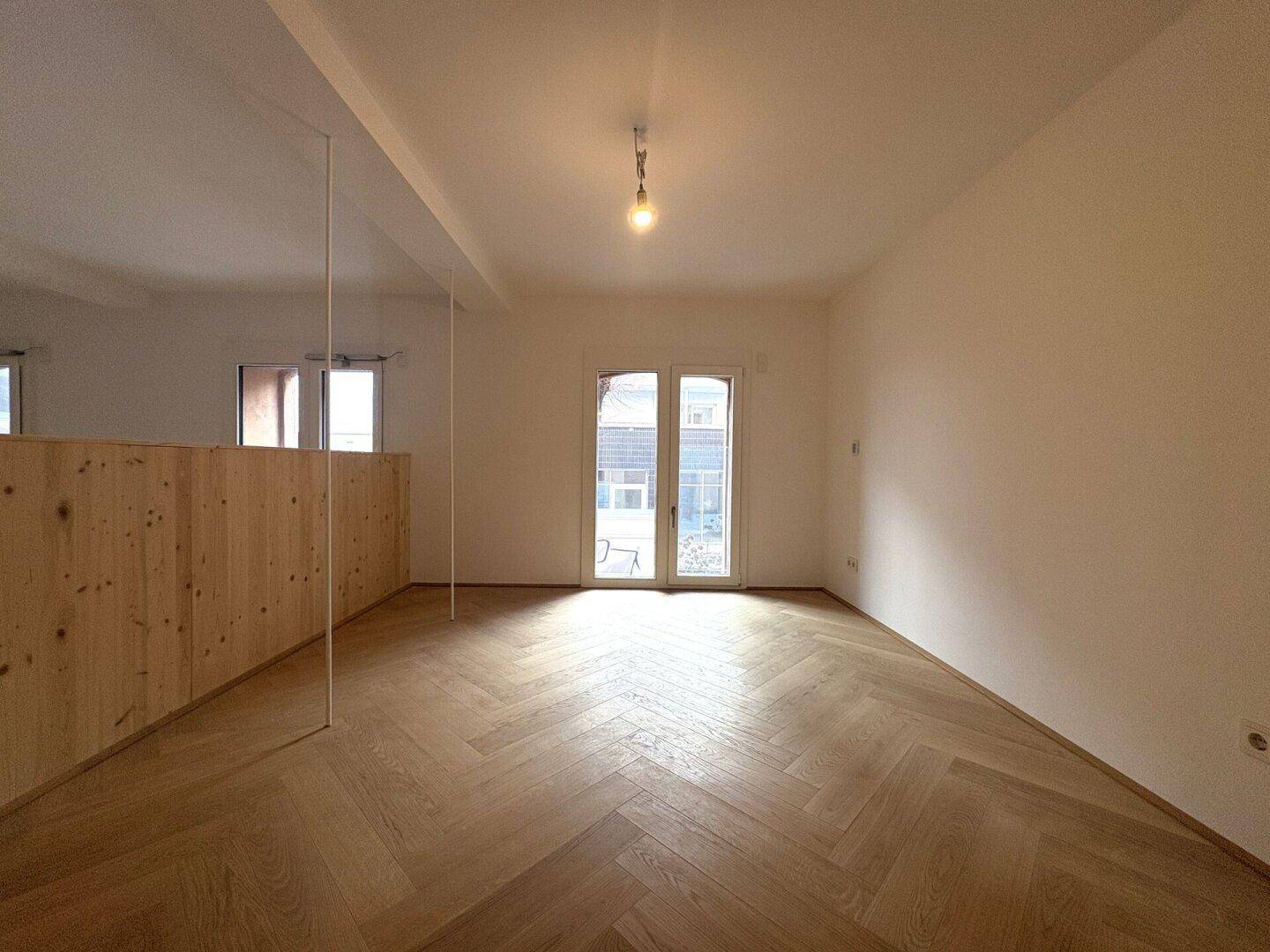 Heller, leerer Raum mit Fischgrätparkettboden, Holzwandverkleidung und Zugang zur Terrasse.