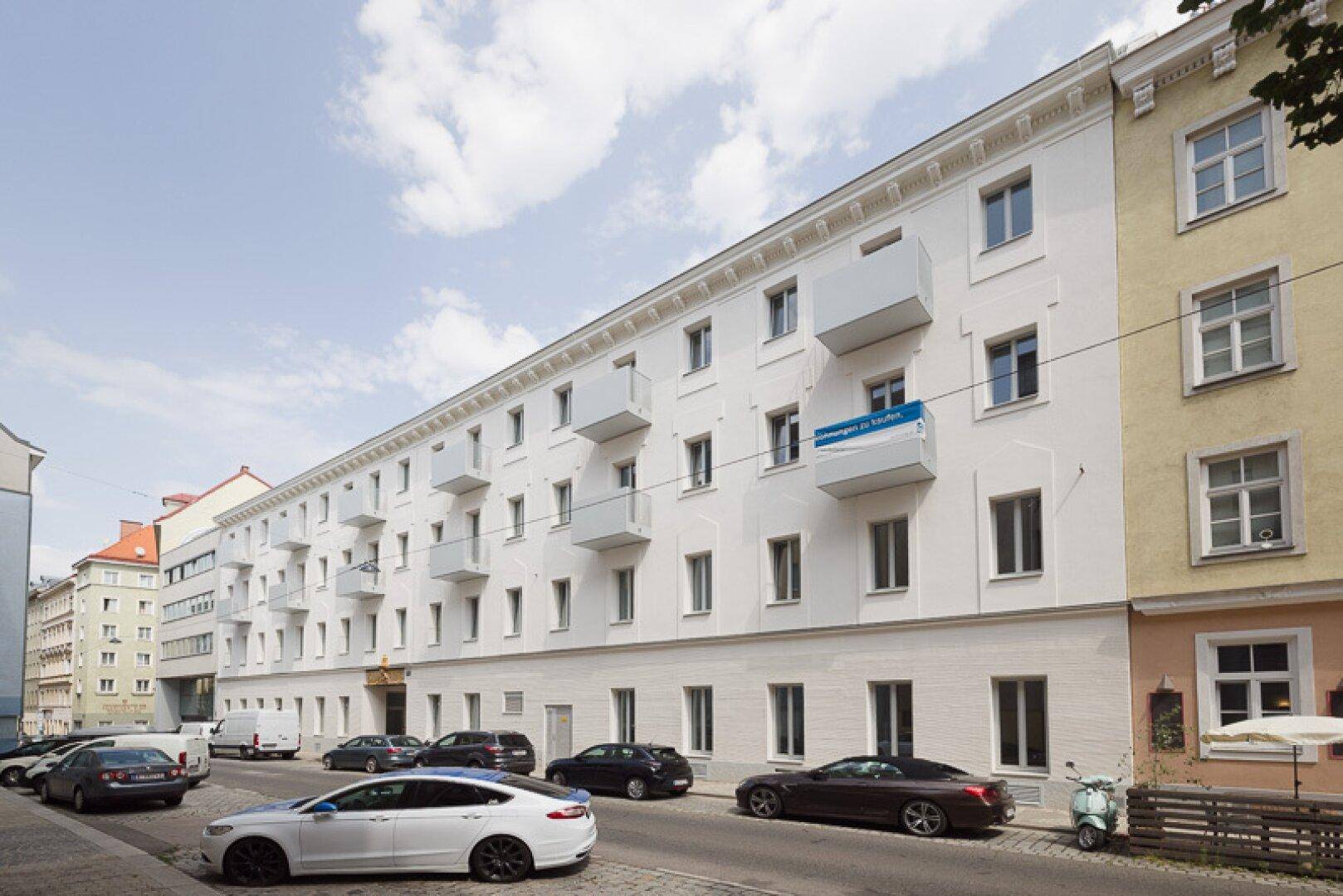 Lange weiße Fassade eines Gebäudes mit vielen Balkonen und parkenden Autos auf der Straße.