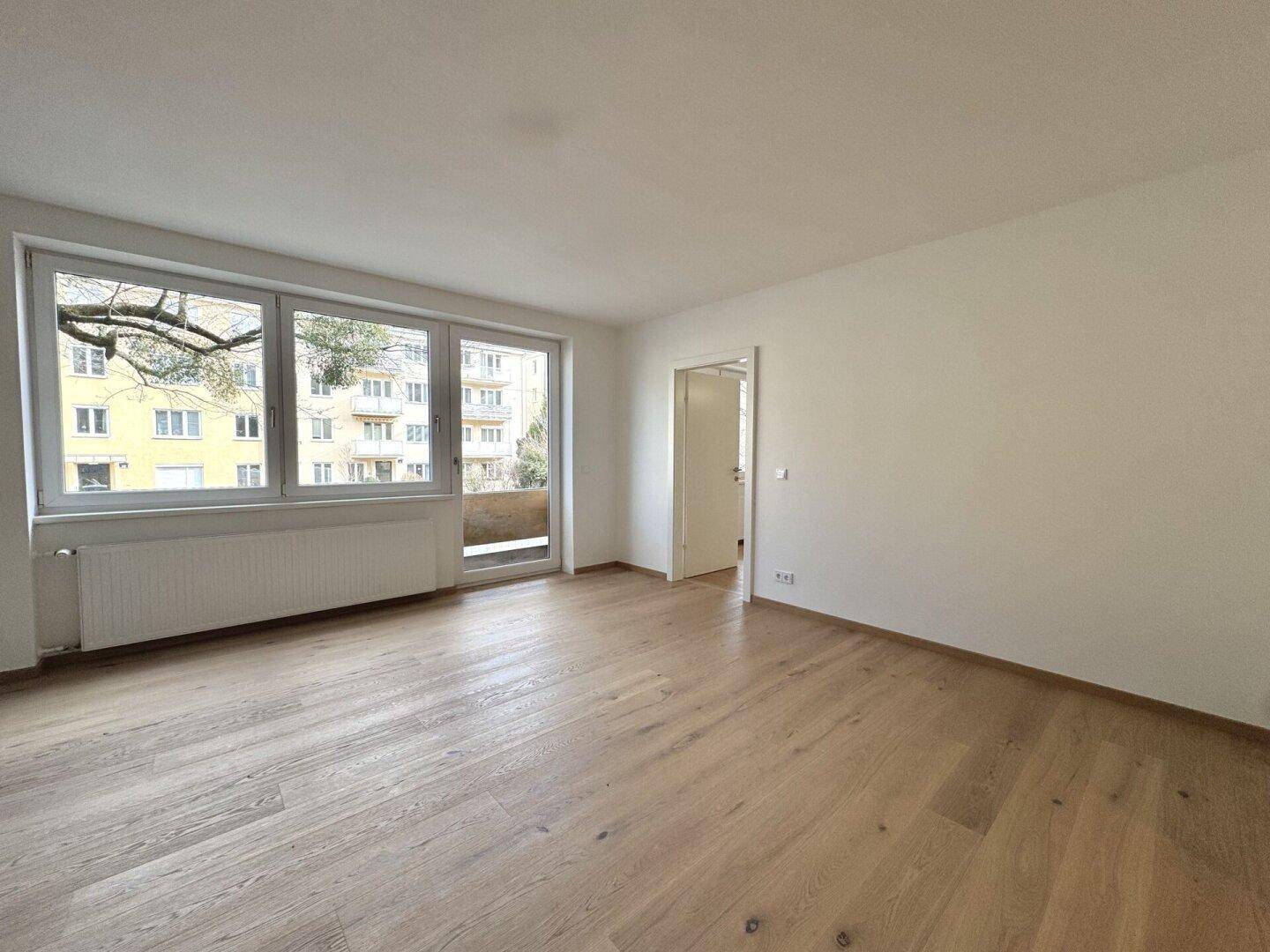 Geräumiges Wohnzimmer mit hellem Parkettboden und großen Fenstern mit Blick nach draußen.