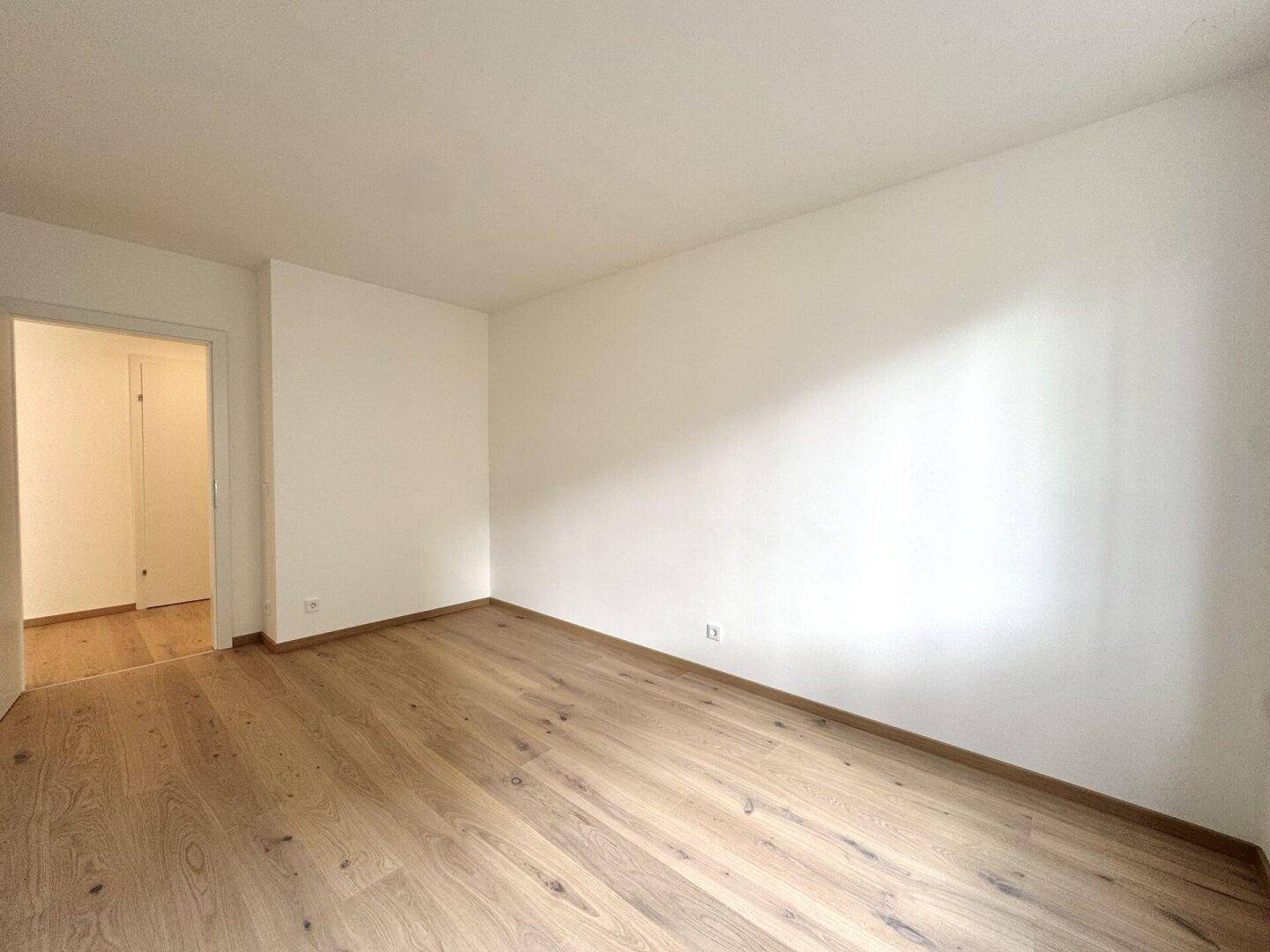 Helles Zimmer mit Holzboden und Zugang zu einem weiteren Raum, mit Sonnenlicht auf dem Boden.
