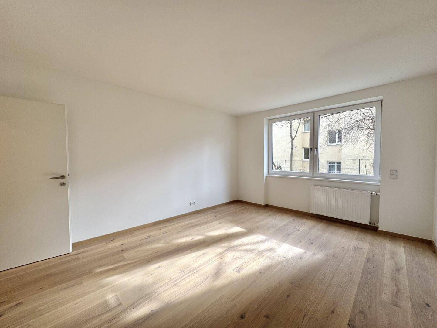 Helles Zimmer mit Holzboden, großem Fenster und einer Tür, die den Raum zugänglich macht.