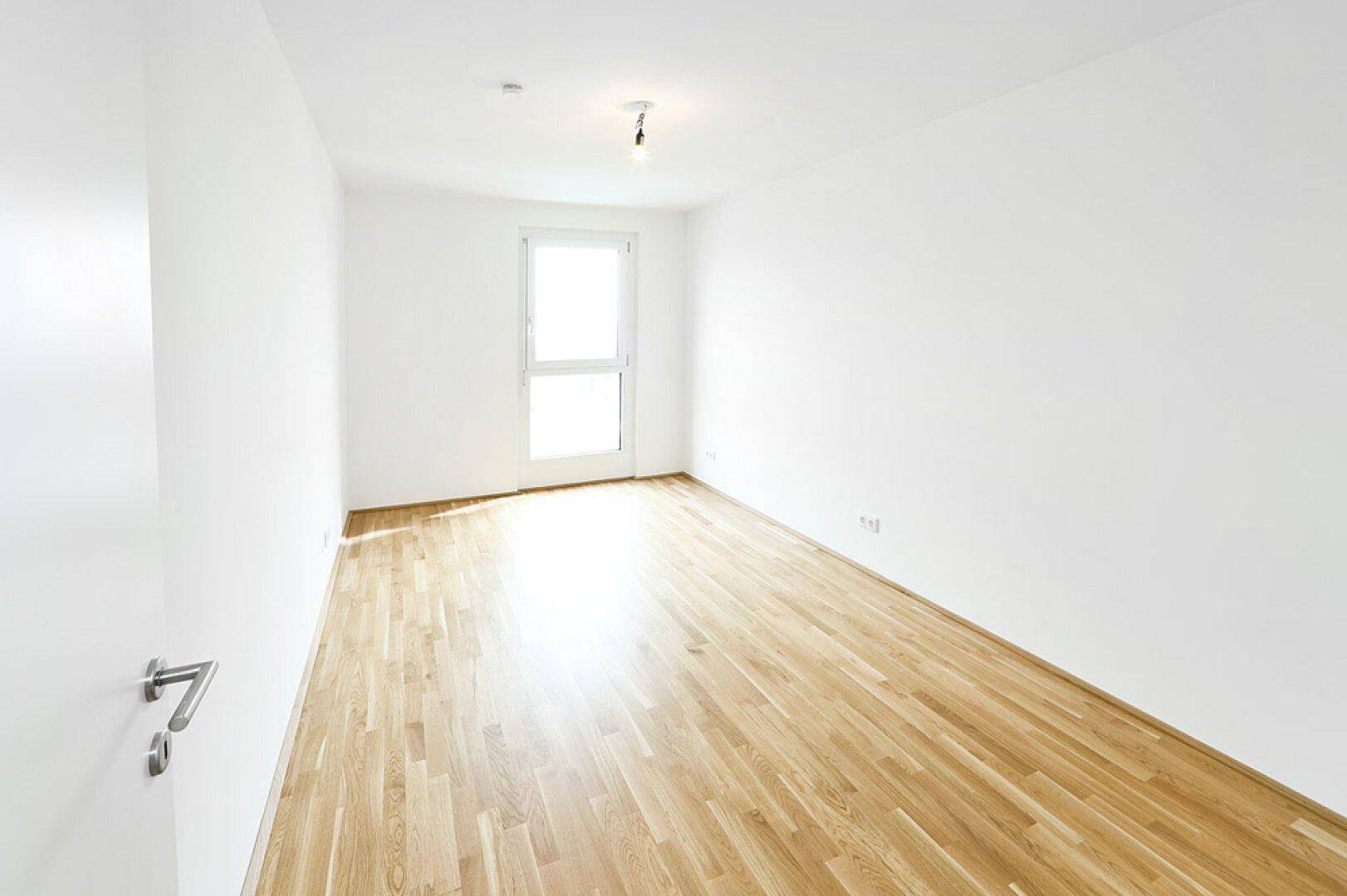Helles Schlafzimmer mit Parkettboden und großem Fenster, das viel Tageslicht hereinlässt.
