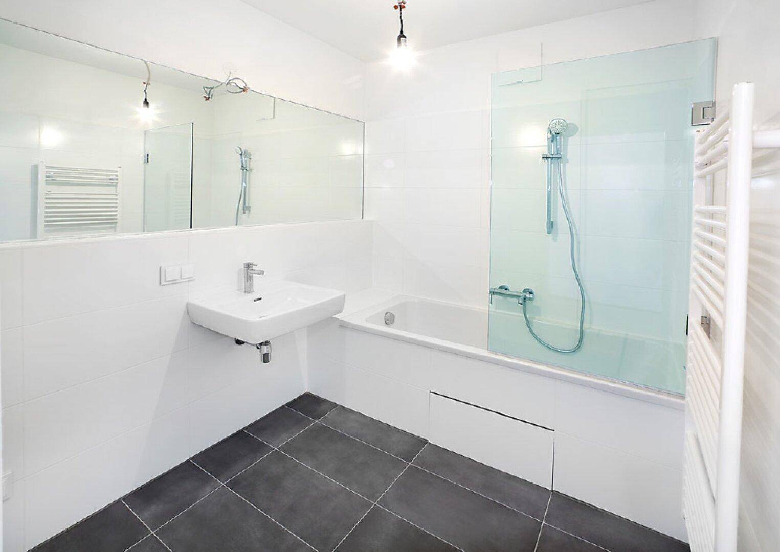 Modernes Badezimmer mit weißen Fliesen, dunklem Boden, Badewanne mit Duschwand und großem Spiegel.