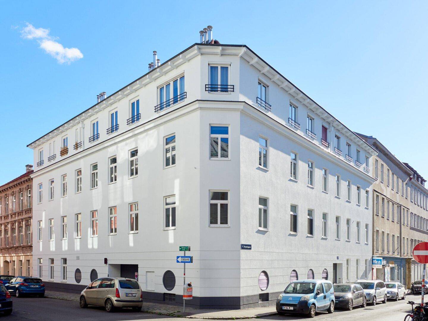 Eckansicht eines frisch renovierten Wohnhauses mit weißer Fassade und vielen Fenstern unter blauem Himmel.