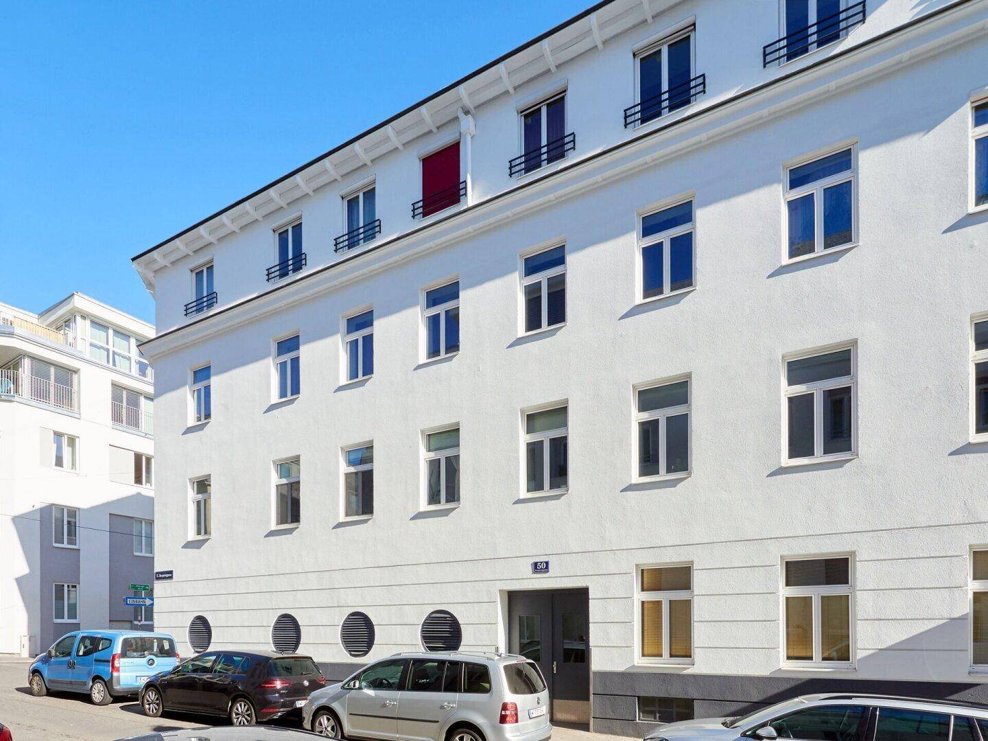 Fassadenansicht des Wohngebäudes mit Eingangsbereich und geparkten Autos auf der Straße.