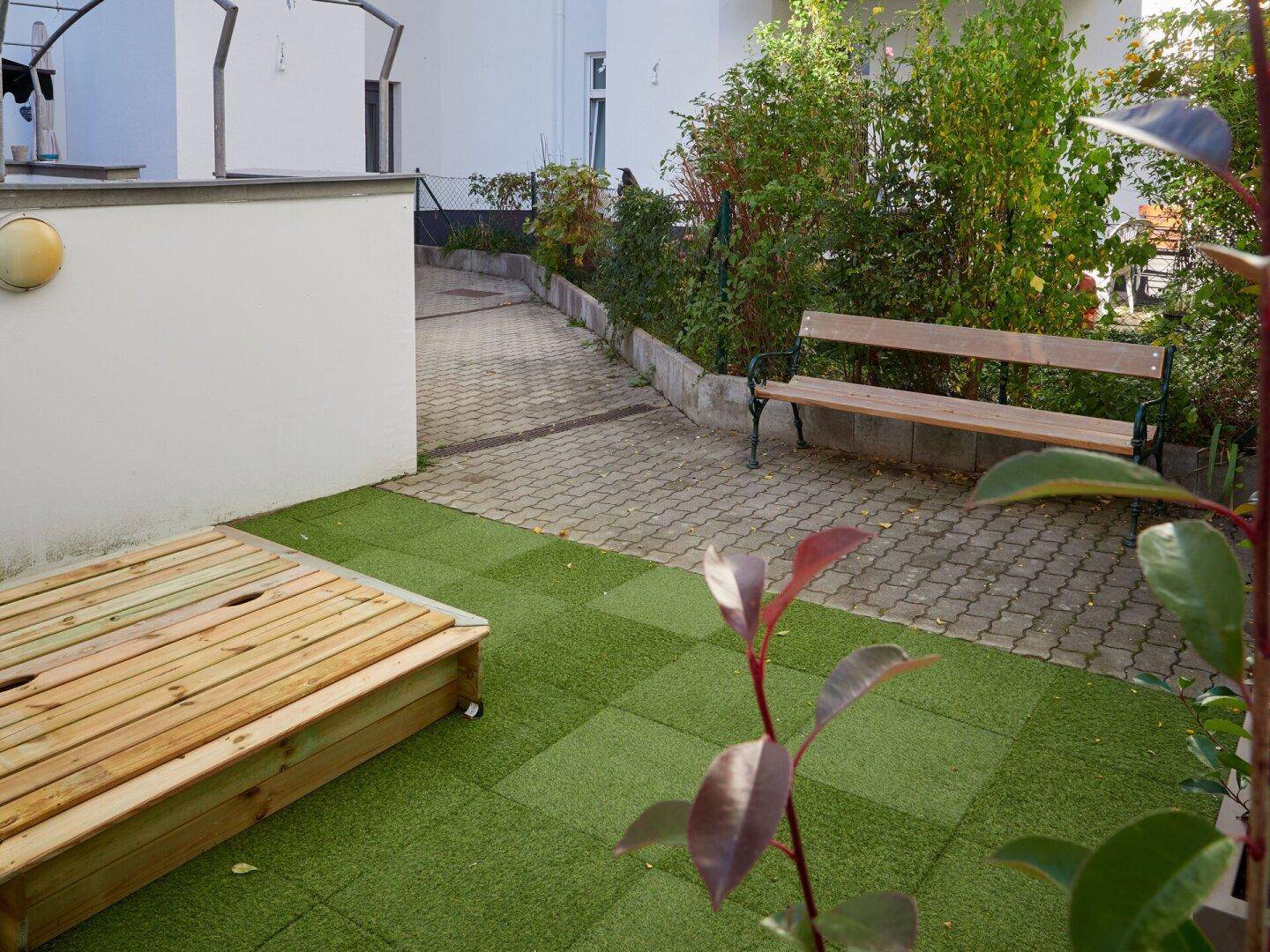 Innenhofbereich mit Sandkasten, Kunstrasen und einer Holzbank unter Bäumen.