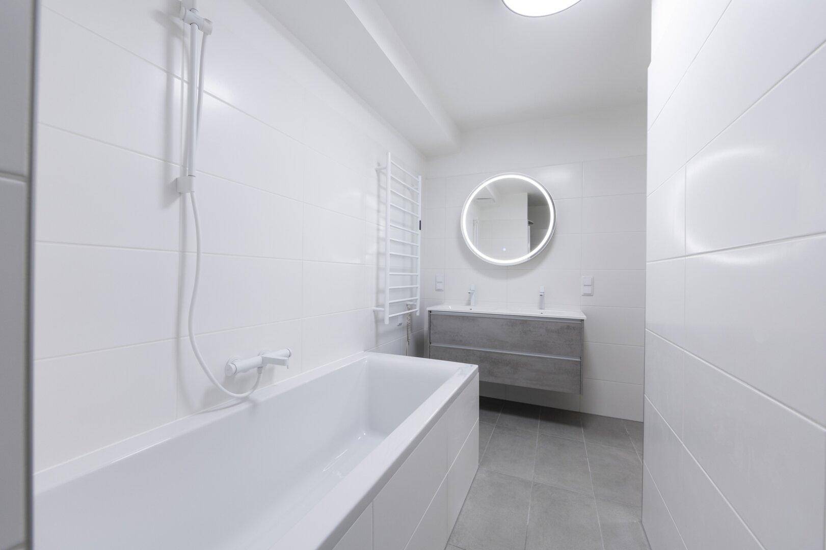 Modernes Badezimmer mit Badewanne, Doppelwaschbecken und beleuchtetem Spiegel in einem hellen, zeitgemäßen Stil.