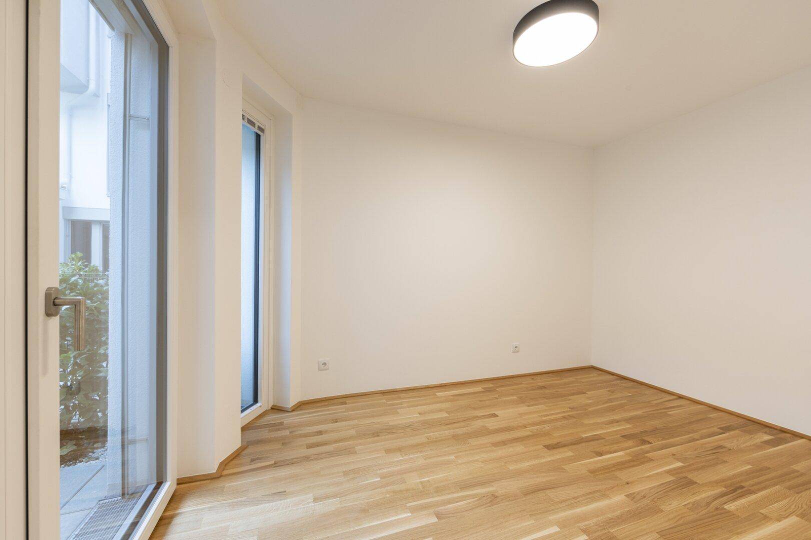 Helles Zimmer mit Parkettboden und bodentiefen Fenstern, die viel Tageslicht hereinlassen und Zugang zum Außenbereich bieten.