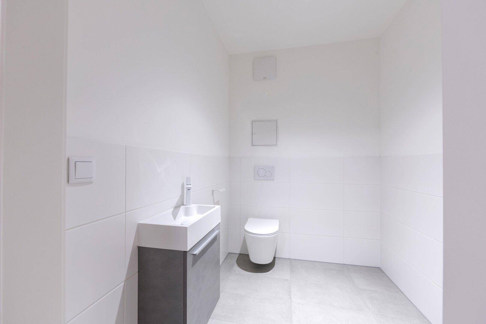 Modernes Gäste-WC mit wandhängender Toilette, kleinem Waschbecken und weißen Fliesen in minimalistischem Design.