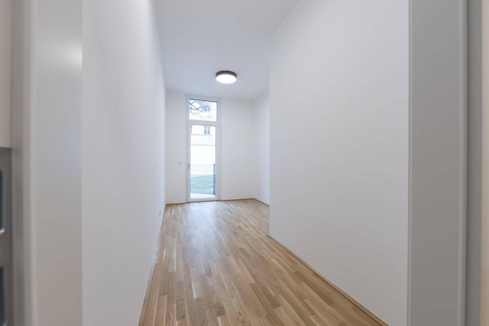 Schmaler, heller Raum mit Parkettboden und einer hohen Fenstertür, die nach draußen in den Garten führt.