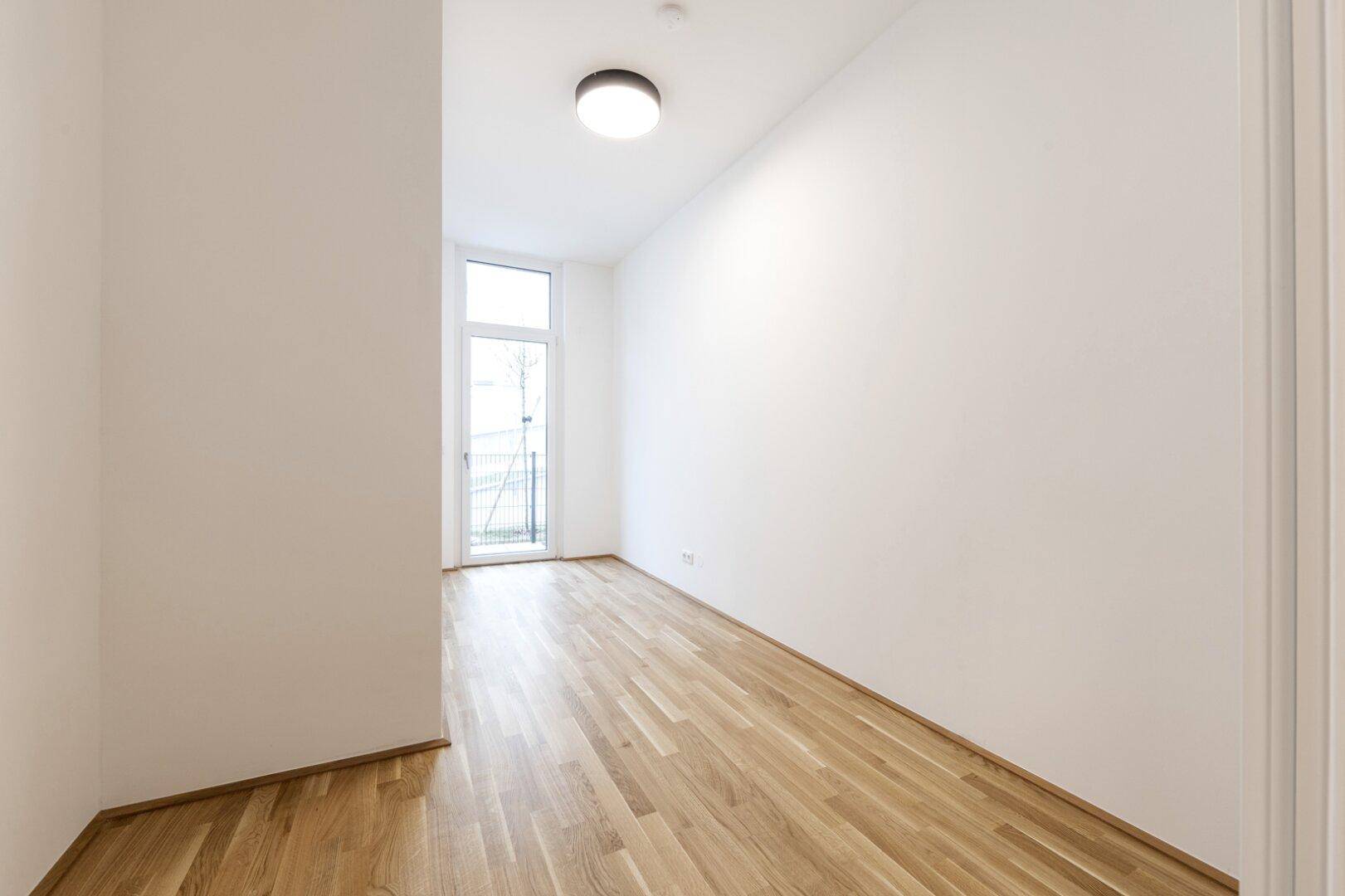 Heller Raum mit Parkettboden und Fenstertür, die zu einem französischen Balkon oder einer kleinen Terrasse führt.