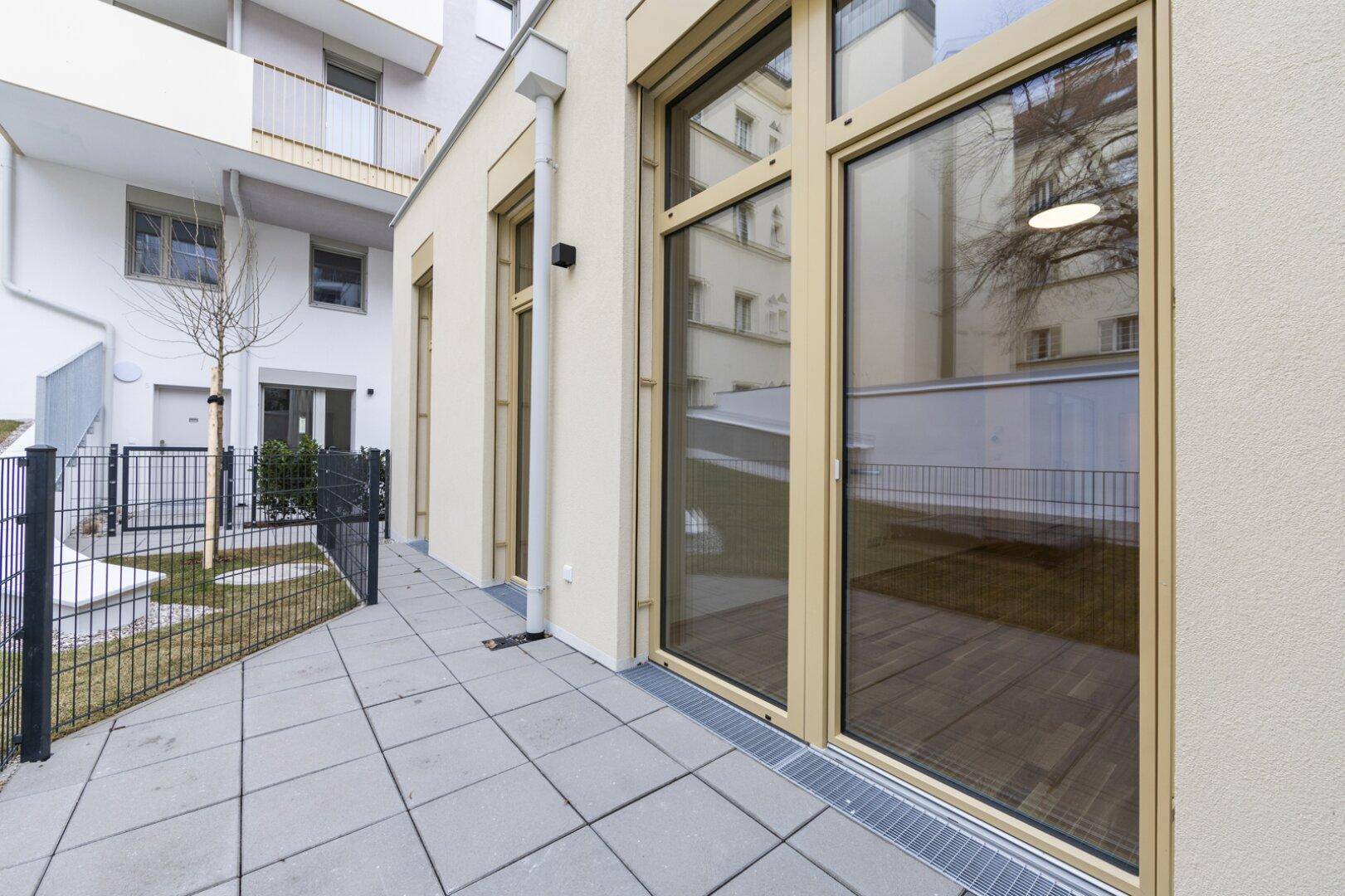 Moderne Terrasse mit Fliesenboden und Zugang zu einem kleinen Gartenbereich, umgeben von Gebäuden.