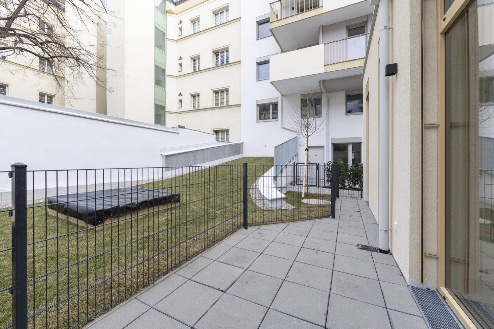 Kleiner privater Gartenbereich mit Rasenfläche und gepflasterter Terrasse, umgeben von einem schützenden Zaun.