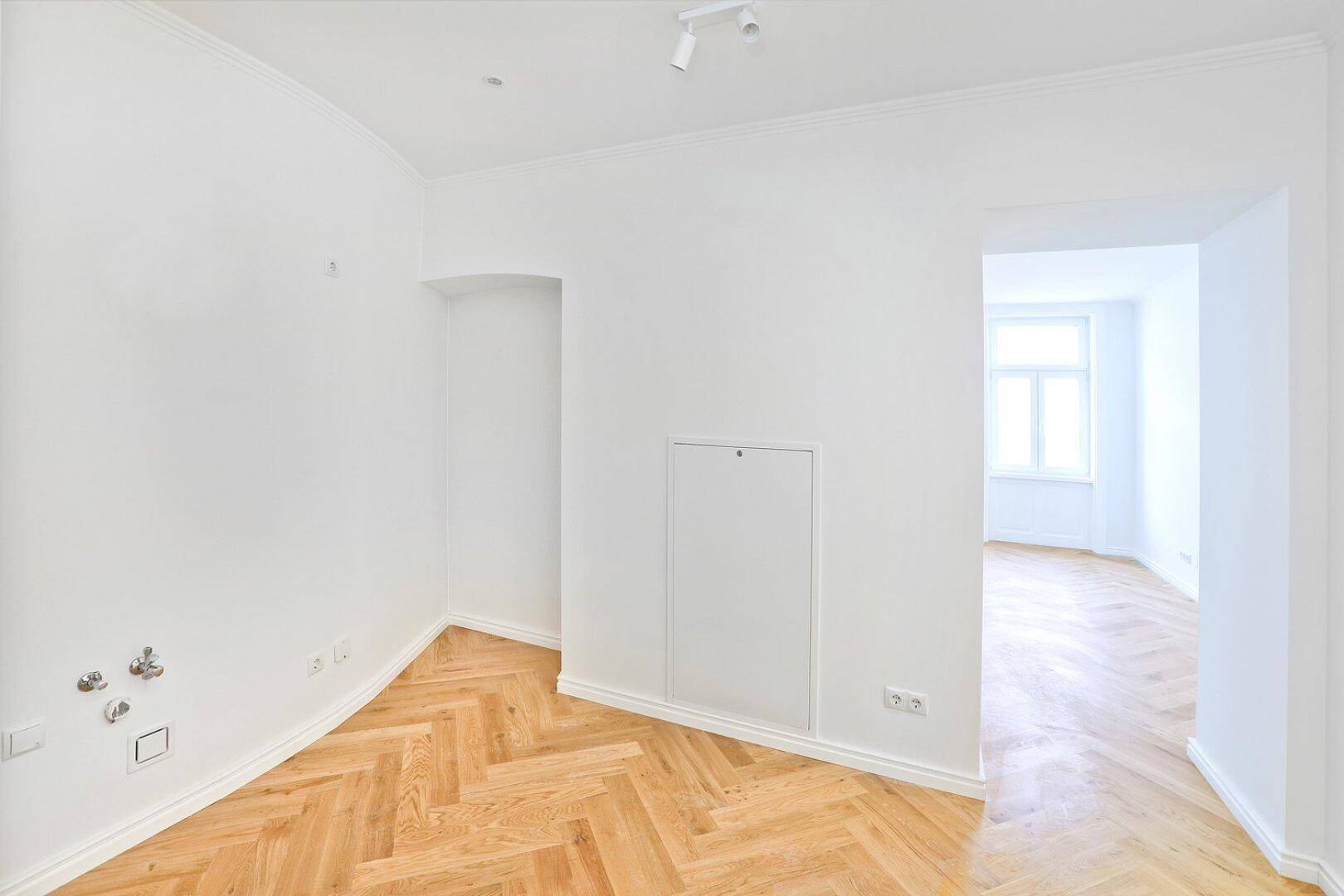 Heller Raum mit Fischgrätparkett und Anschlüssen für eine zukünftige Küche.