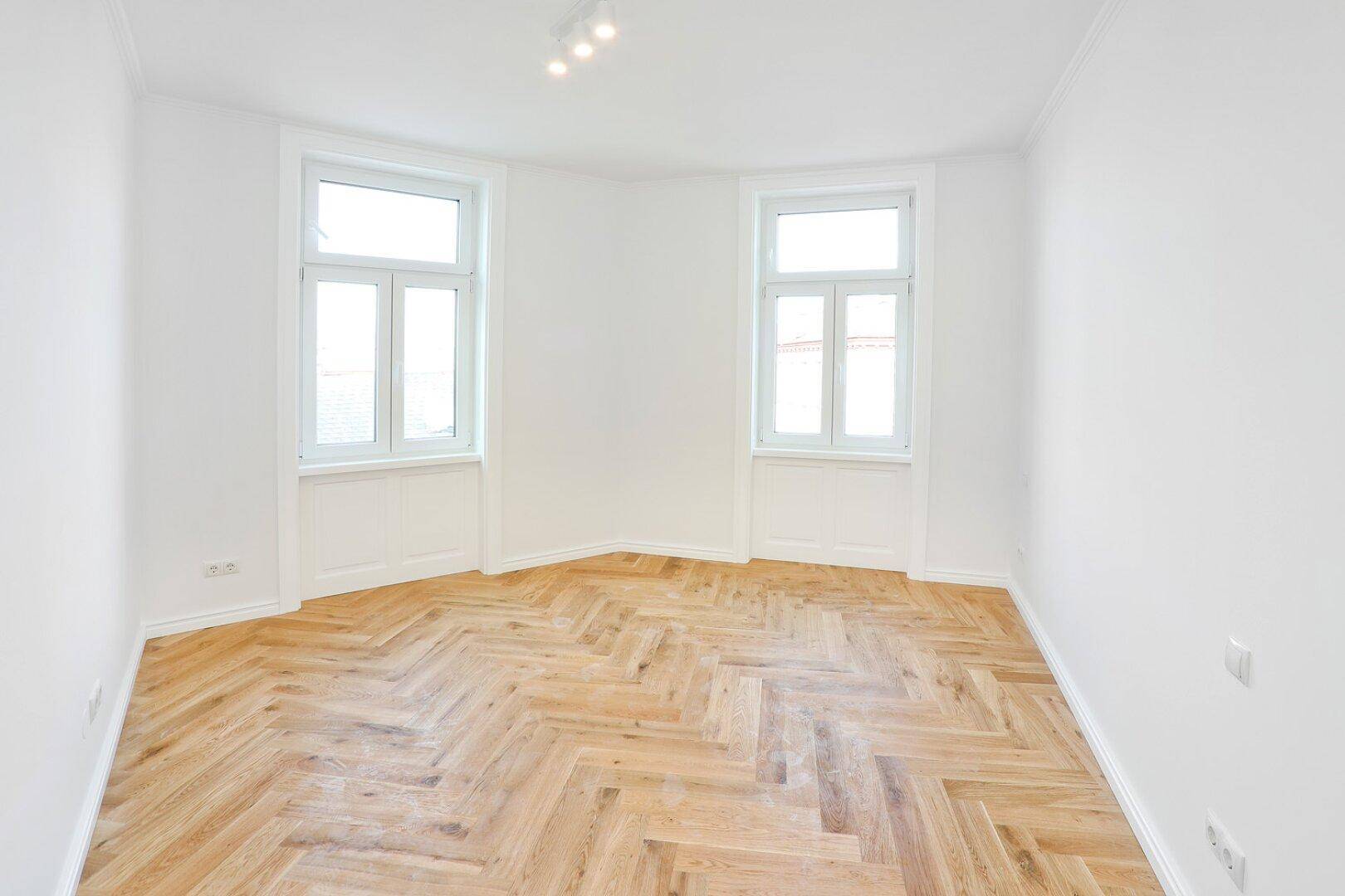 Geräumiges Zimmer mit hellem Fischgrätparkett und zwei Fenstern, die viel Tageslicht hereinlassen.
