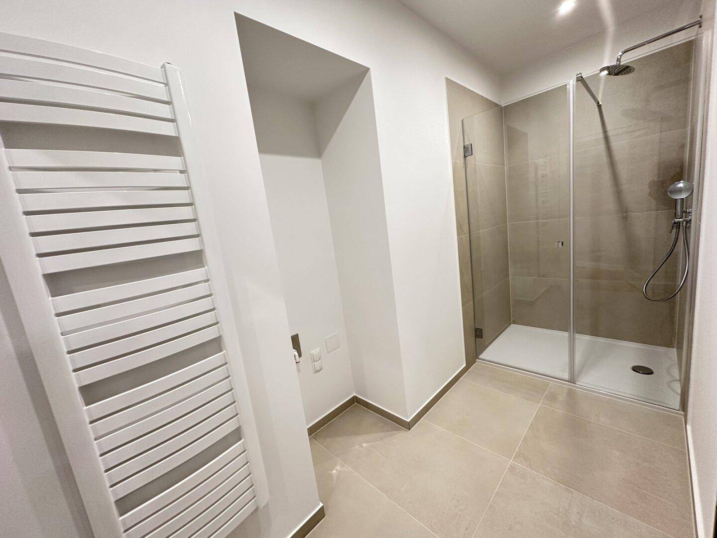 Helles Badezimmer mit großer begehbarer Dusche, neutralen Fliesen und einem praktischen Handtuchheizkörper.