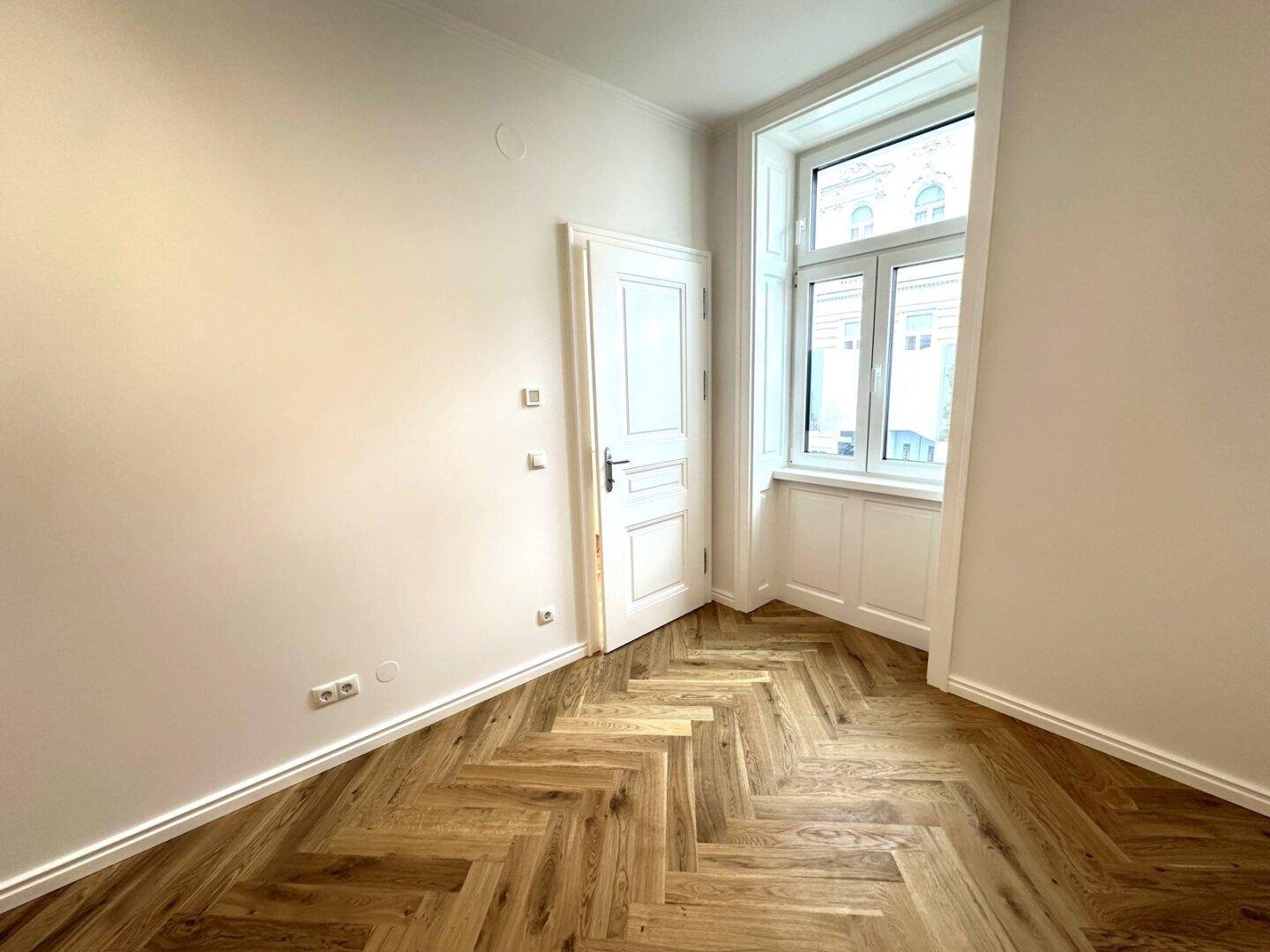 Lichtdurchflutetes Zimmer mit klassischem Fischgrätparkett und einem hohen Fenster, das den Raum erhellt.