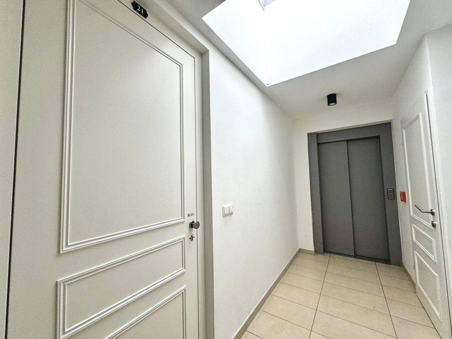 Heller Flur mit weißer Wohnungstür, Aufzug und einem Oberlicht, das Tageslicht hereinlässt.