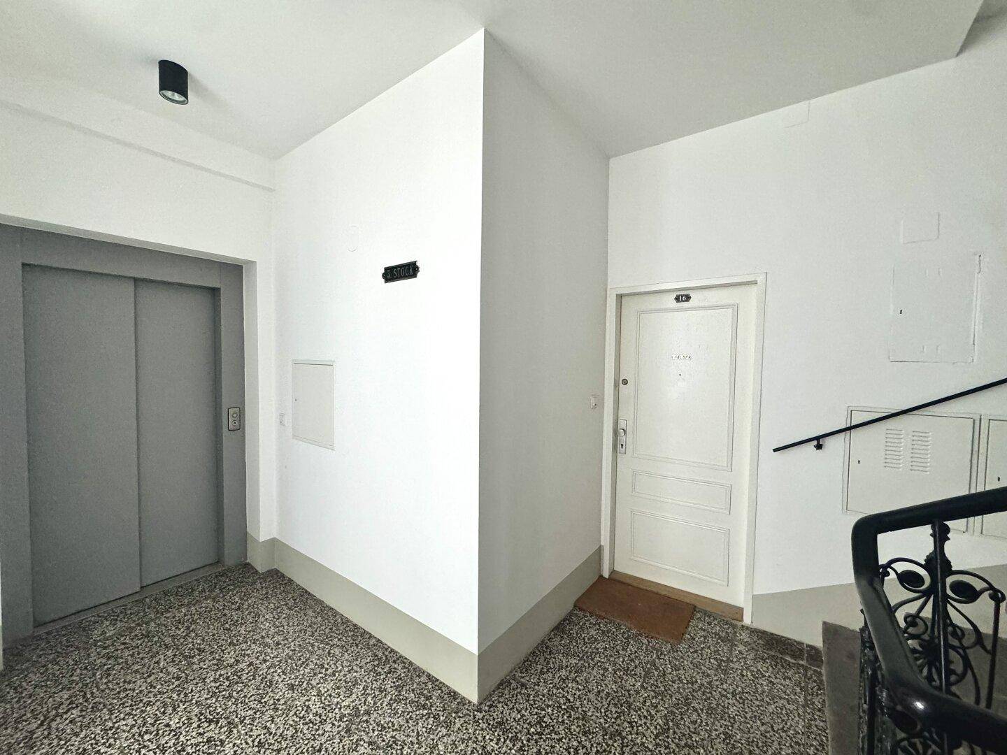 Eingangsbereich mit Aufzug, einer weißen Wohnungstür und Treppenaufgang mit dunklem Geländer.